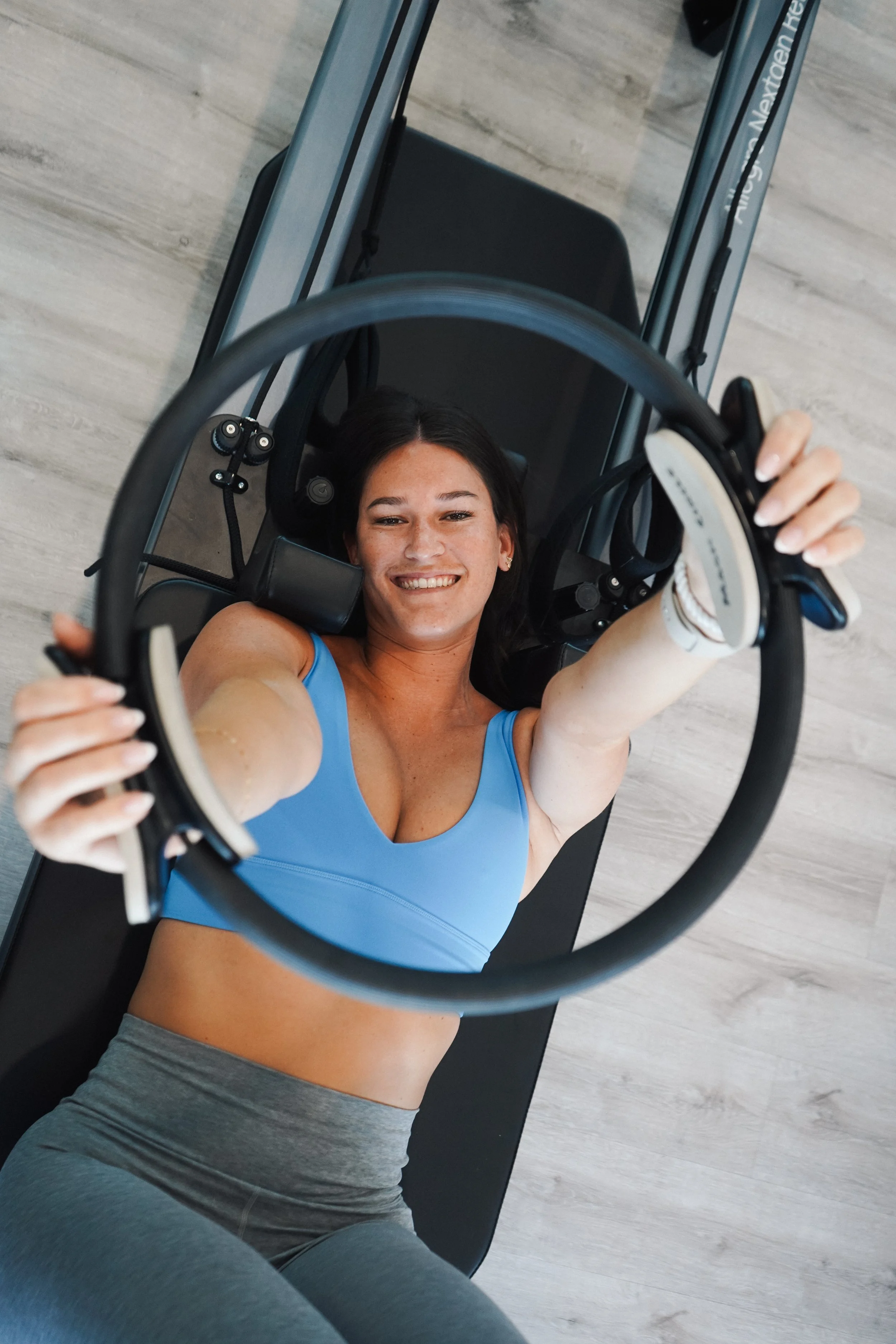 Woman lying on her back on a black exercise mat, smiling, holding a Pilates ring above her head, wearing a blue sports bra and gray leggings.