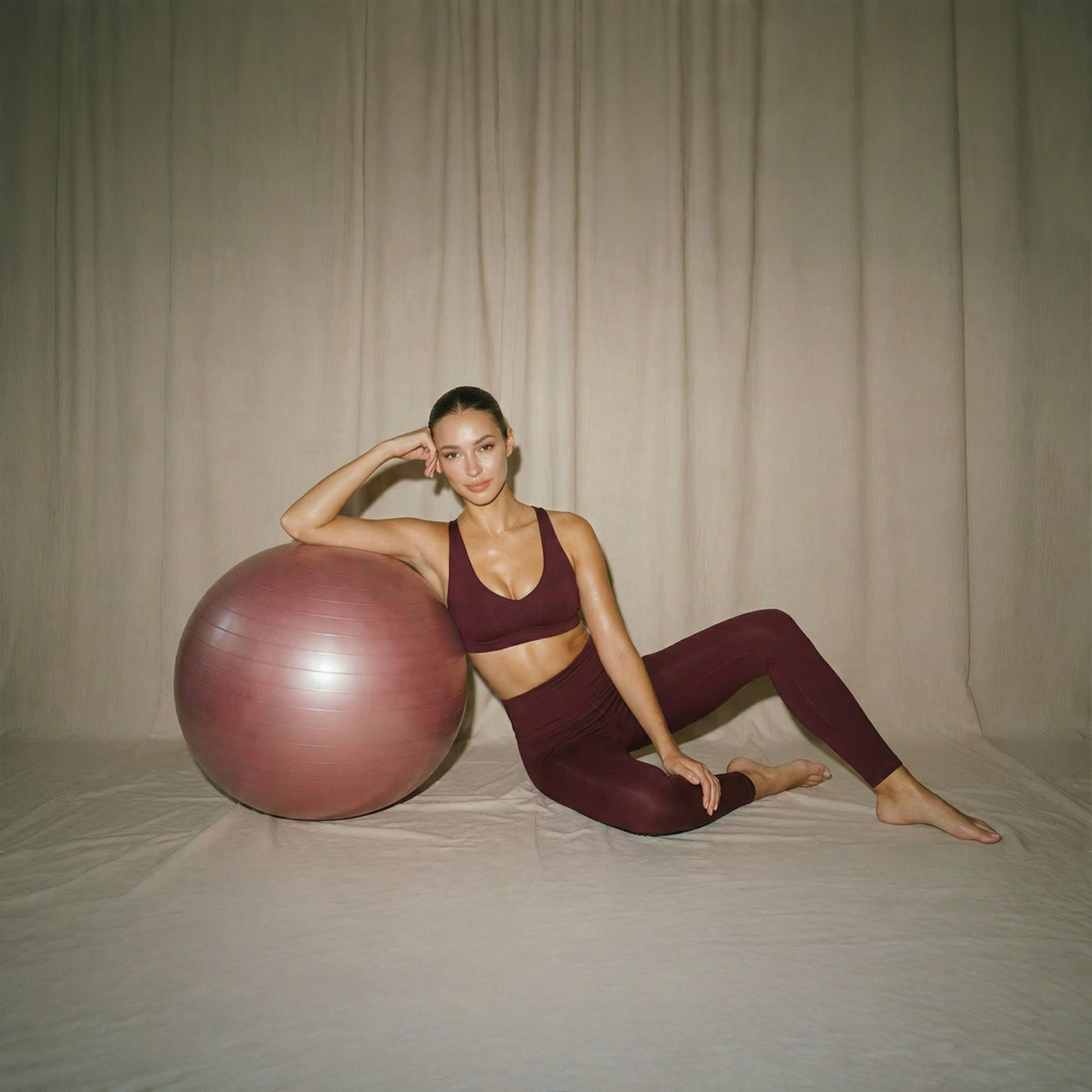 A woman in activewear sitting on the floor, resting her head on her hand, with a large pink exercise ball in front of her.