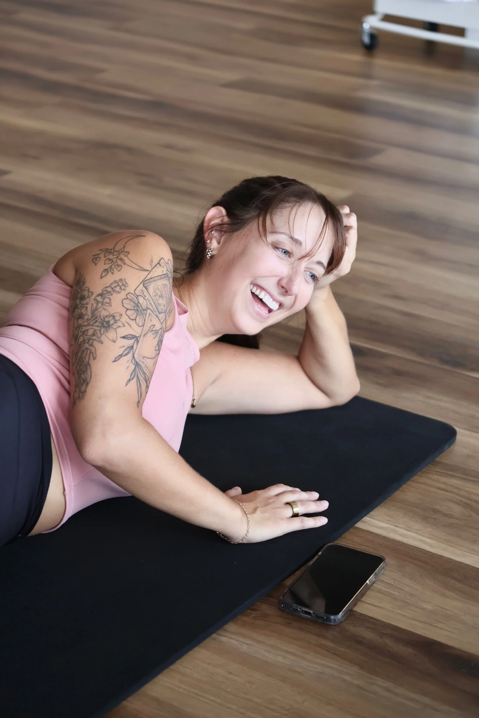 A woman lying on a black yoga mat on a wooden floor, smiling and holding her head with one hand. She has short brown hair, multiple earrings, and a tattoo sleeve on her left arm. A smartphone is on the floor near the yoga mat.