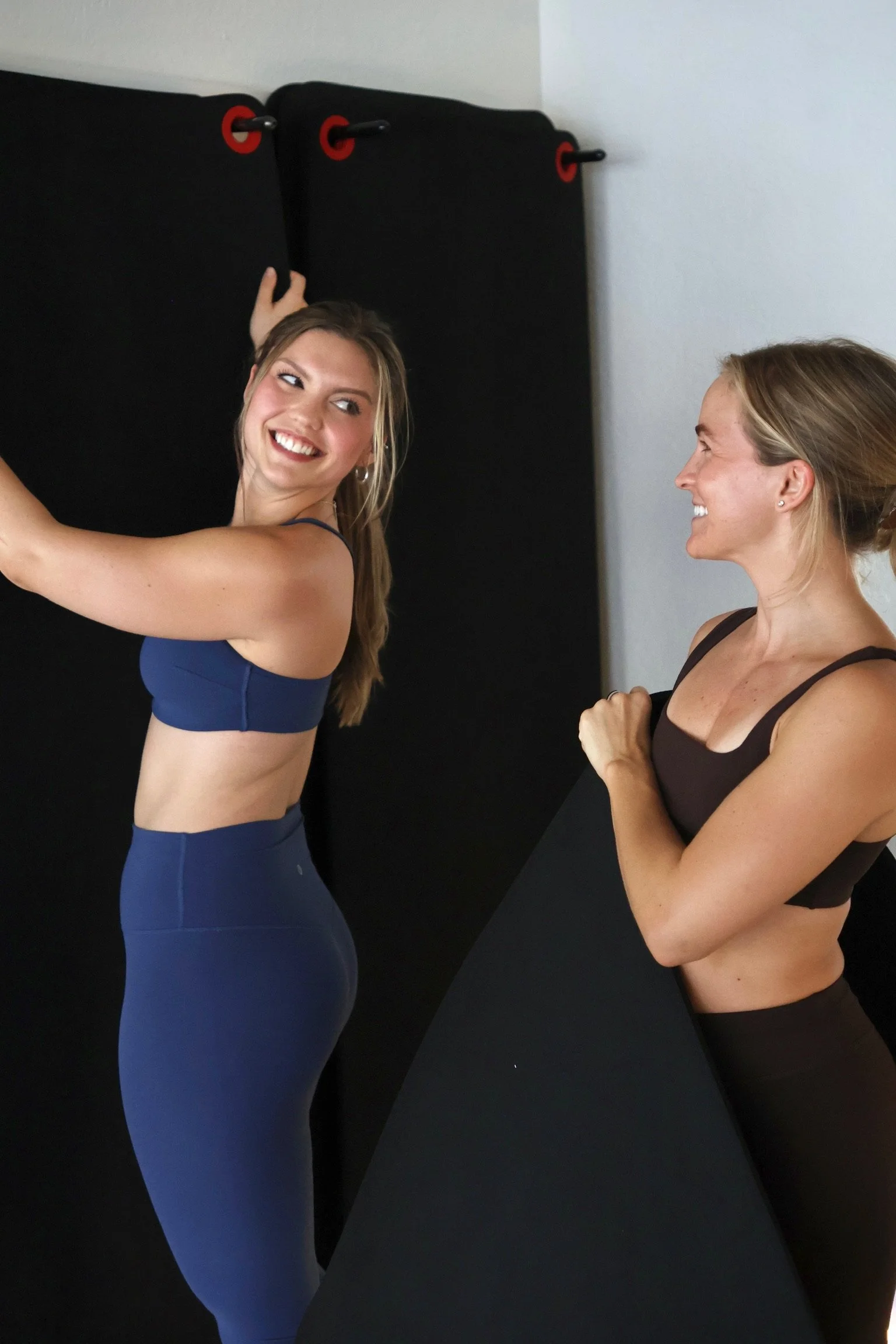 Two women in workout clothes smiling in a fitness studio, one adjusting a black curtain or backdrop on a black wall.