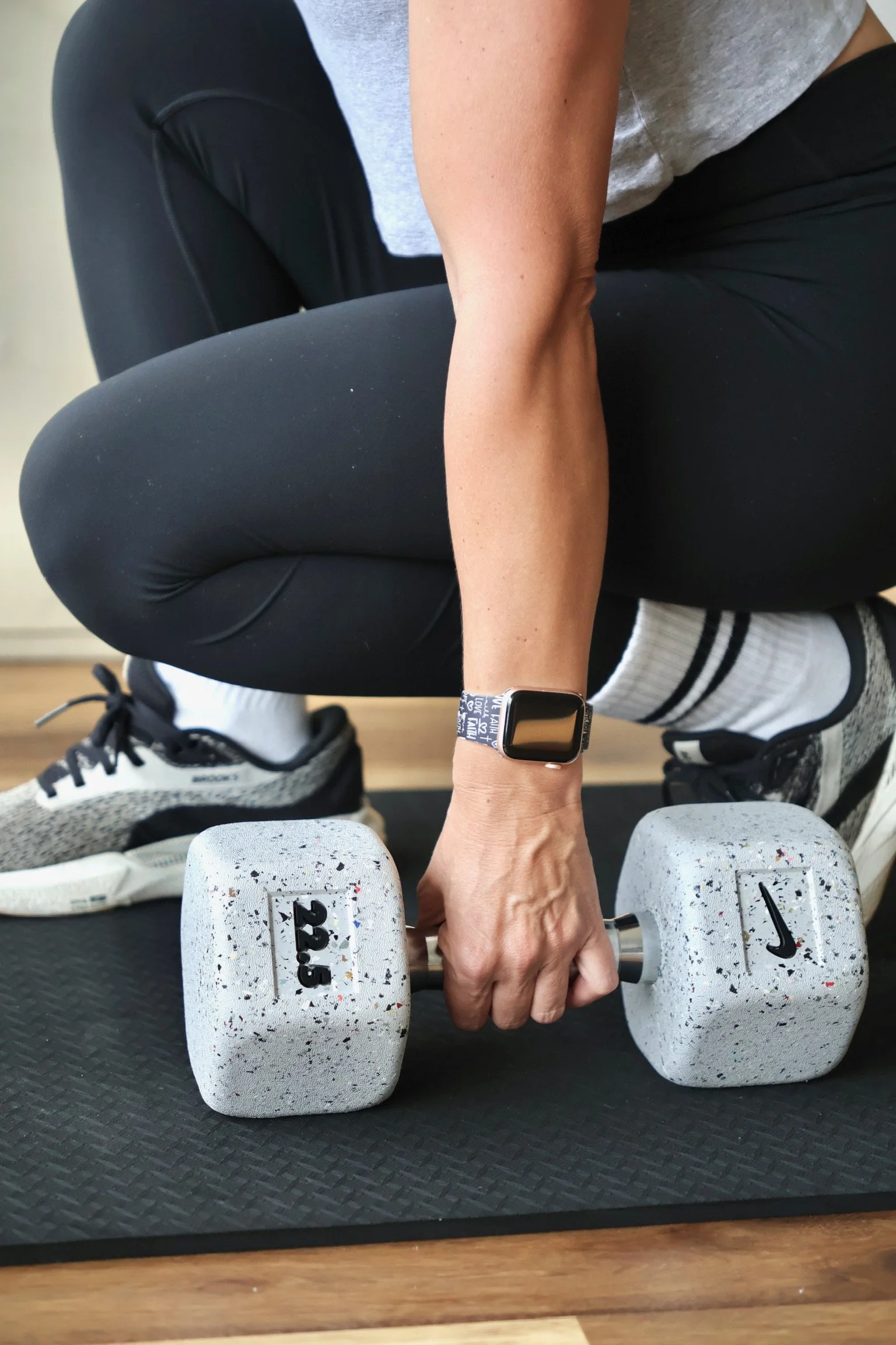 Person doing a workout with dumbbells on a black exercise mat, wearing a smartwatch, gray t-shirt, black leggings, and white sneakers.