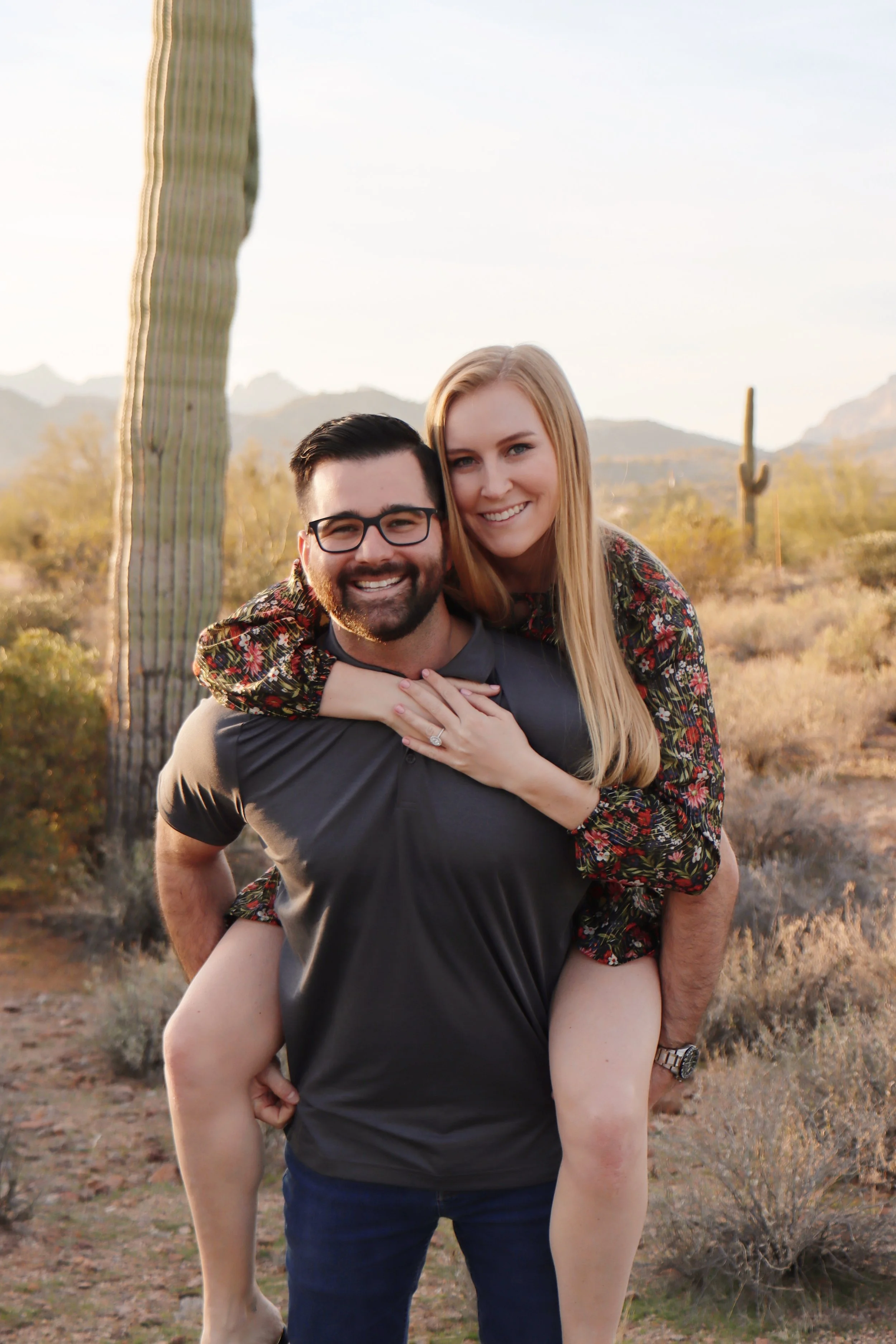 A smiling man with glasses carrying a smiling woman with long blonde hair on his back in a desert landscape with cacti and mountains in the background.