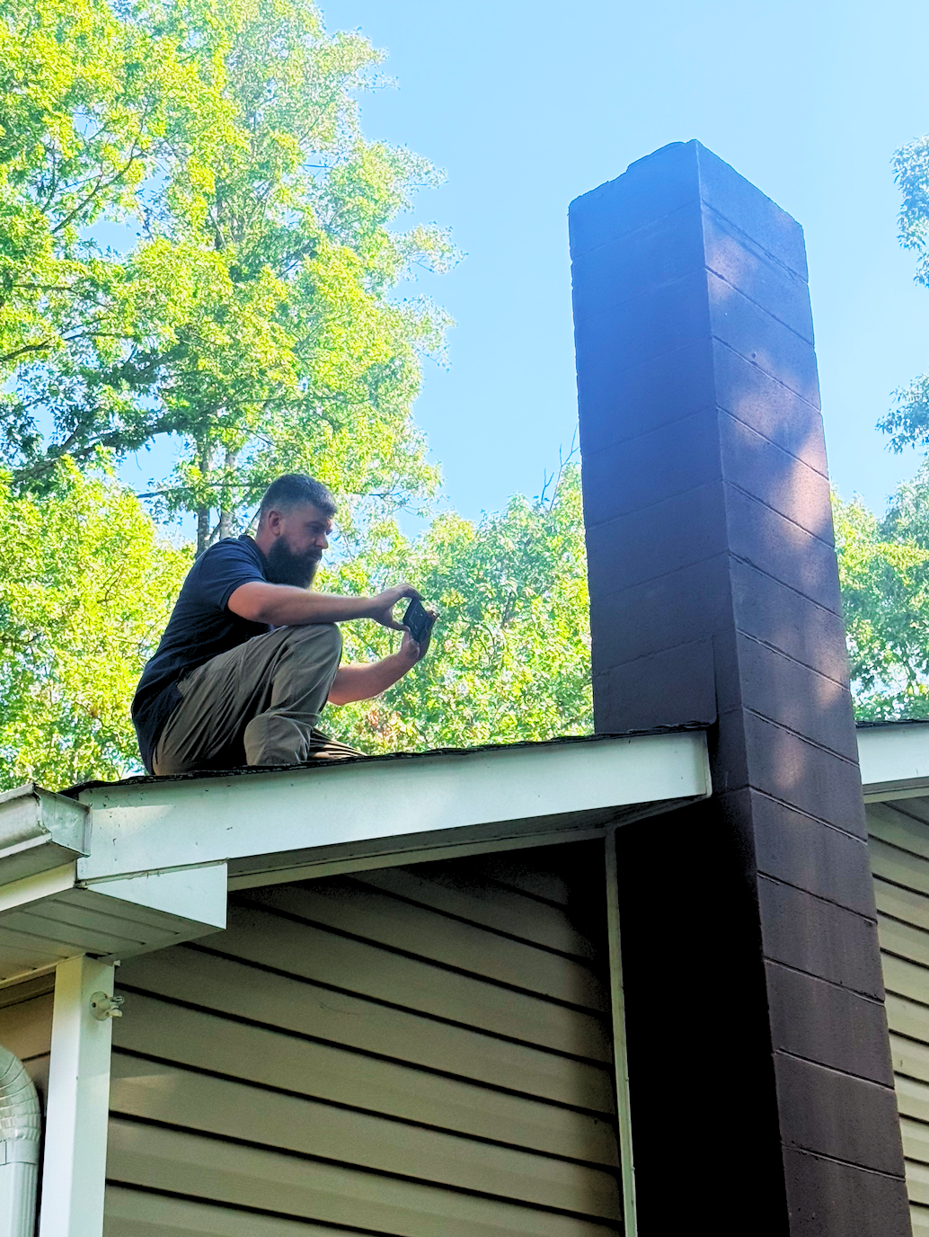 Man (Jeff) sitting on the roof of a house taking a photo of the chimney with a smartphone, surrounded by green trees and a clear blue sky.