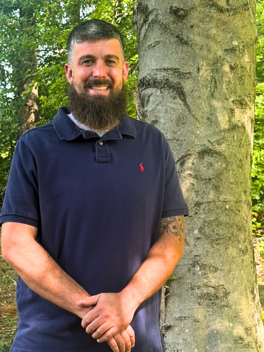 A smiling man (Jeff) with a beard and short hair standing outdoors next to a large tree with textured bark, surrounded by green foliage.