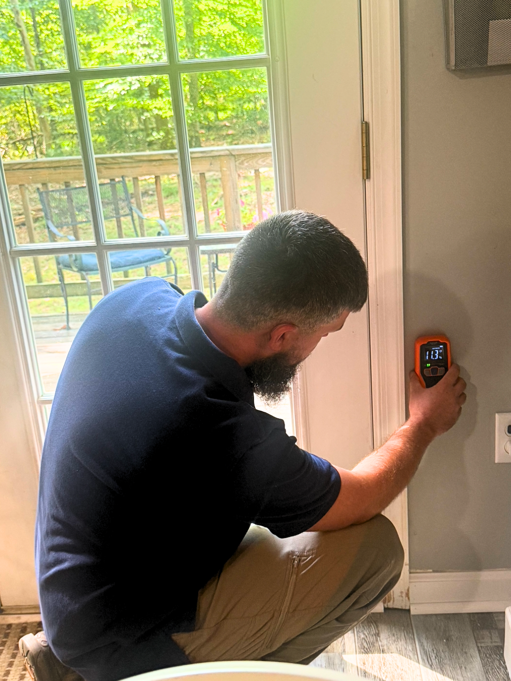 A man with dark hair (Jeff) and a beard kneeling by a window, using a digital thermometer on the wall.