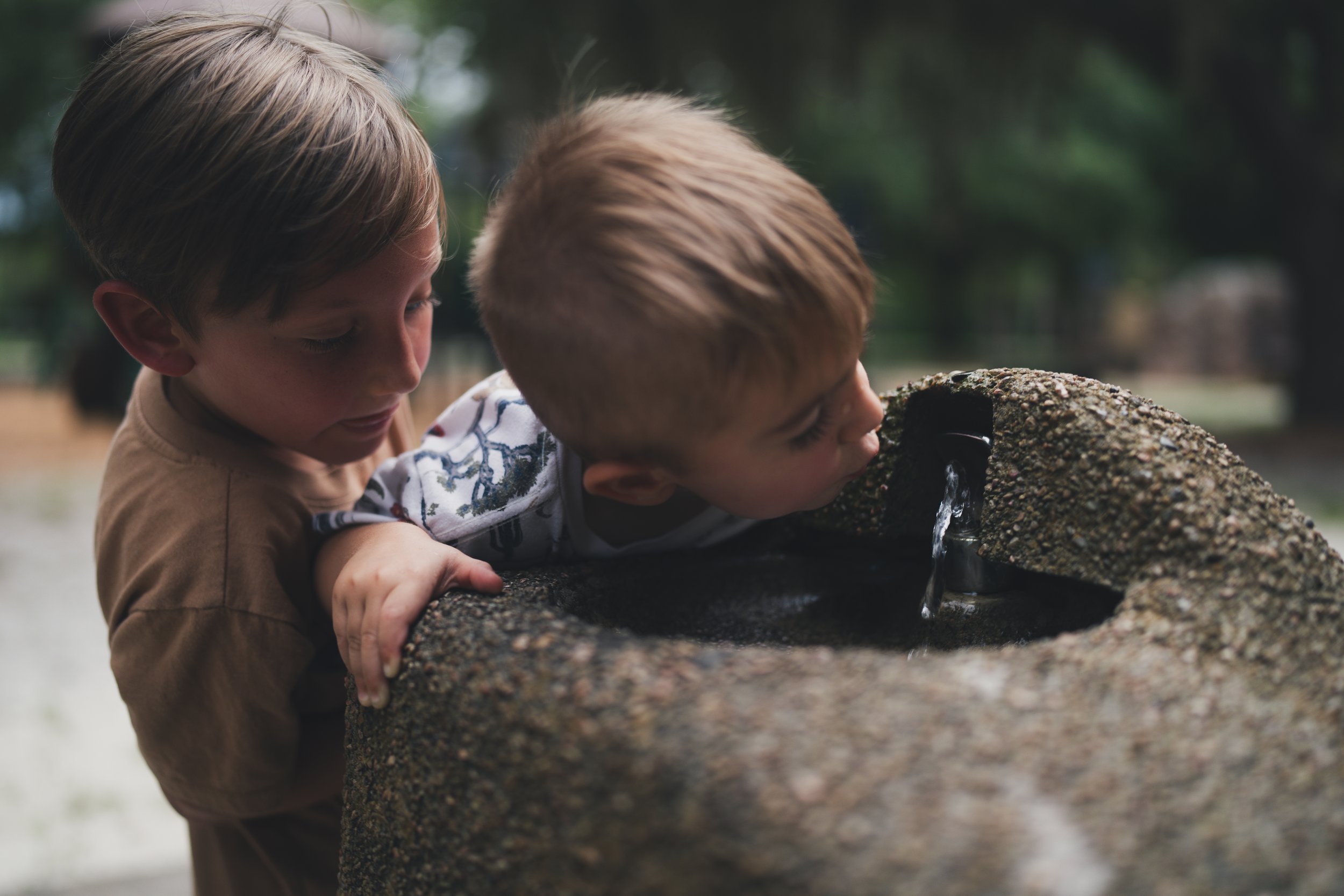 Two young boys leaning over a stone water fountain outdoors, with one boy drinking water from the fountain.