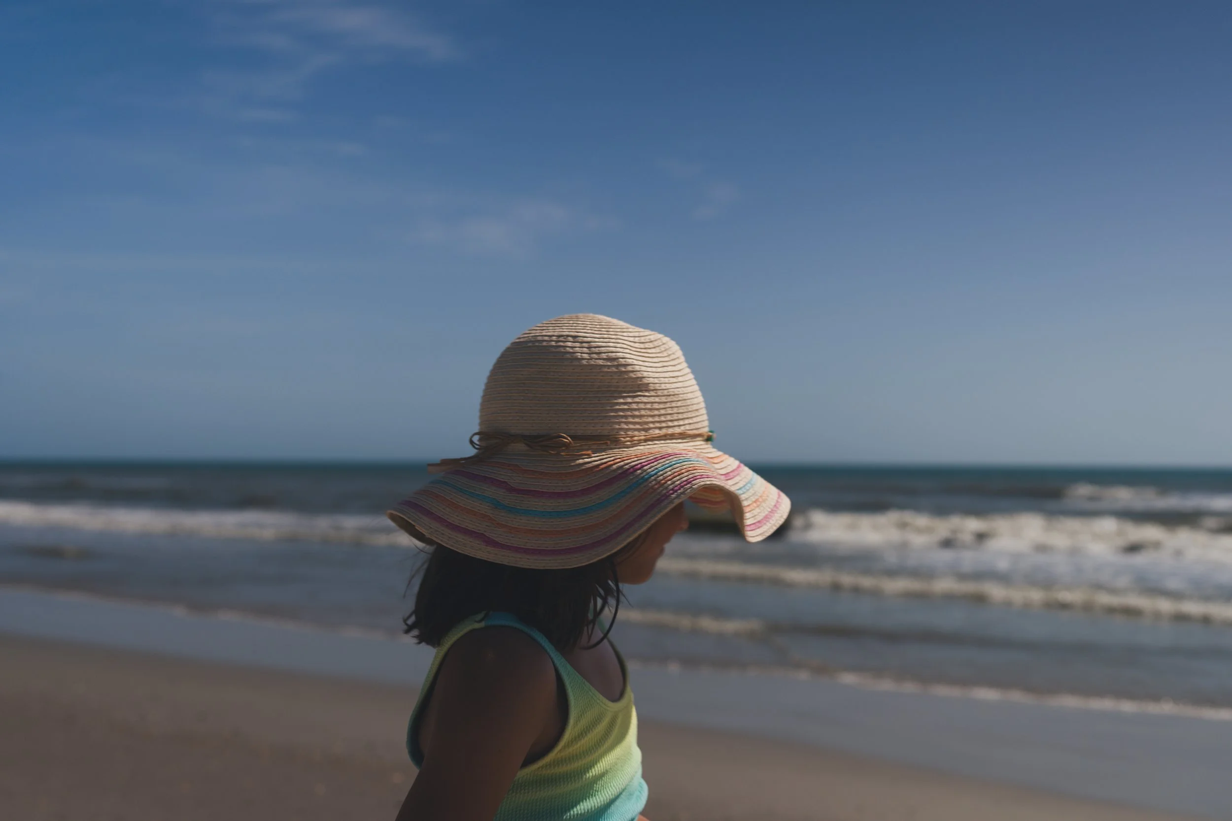 A young girl wearing a colorful wide-brimmed hat and a bright yellow swimsuit walking along the beach shoreline with waves in the background under a clear blue sky.