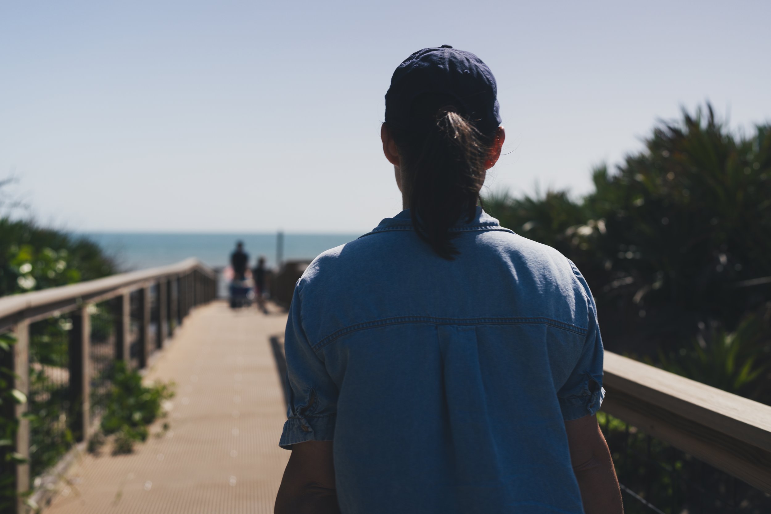 Person wearing a dark hat and a denim jacket walking on a wooden boardwalk along the coast with a view of the ocean and some blurred people in the background.