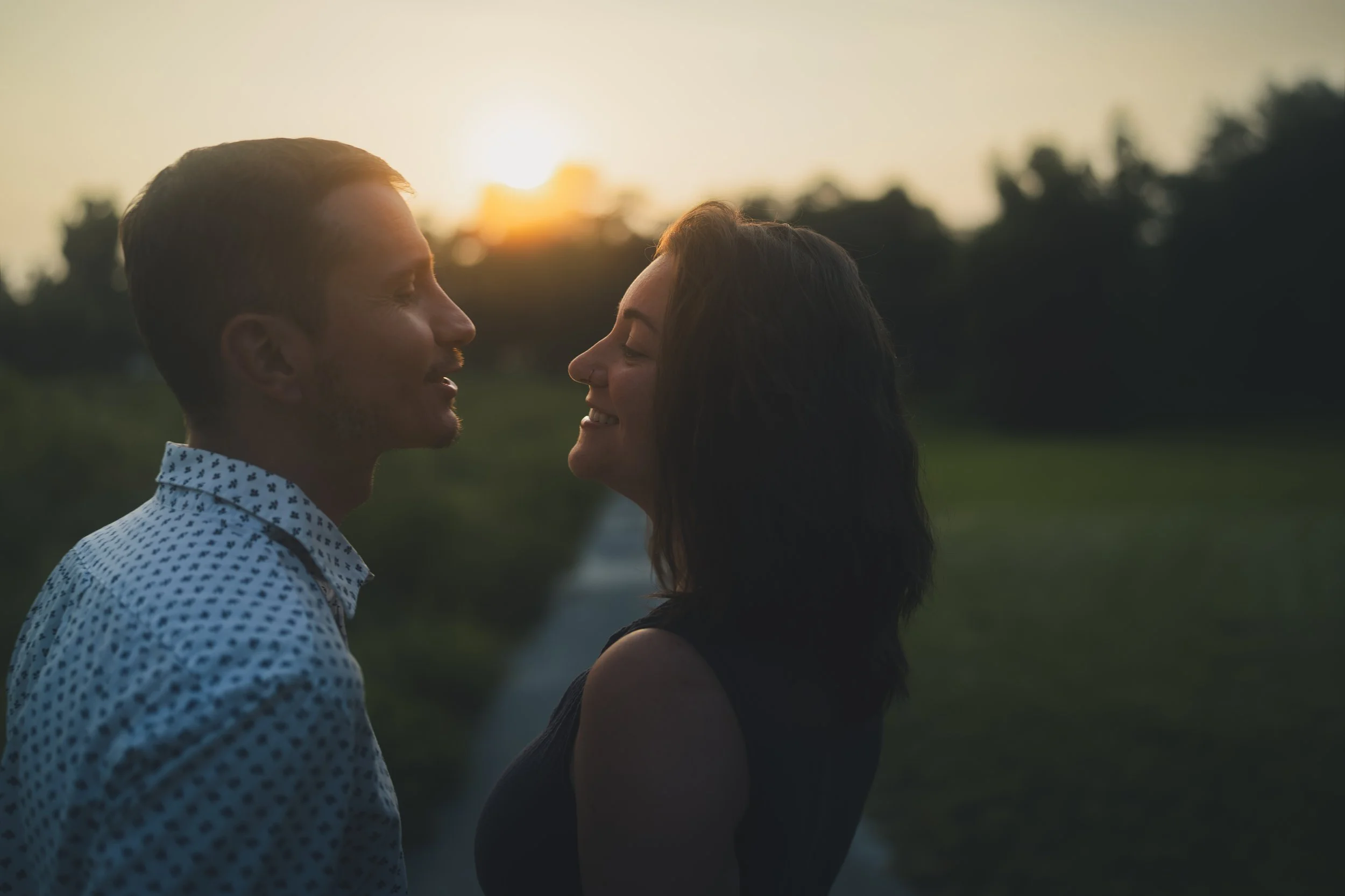 A couple standing close together outdoors during sunset, facing each other with smiles.
