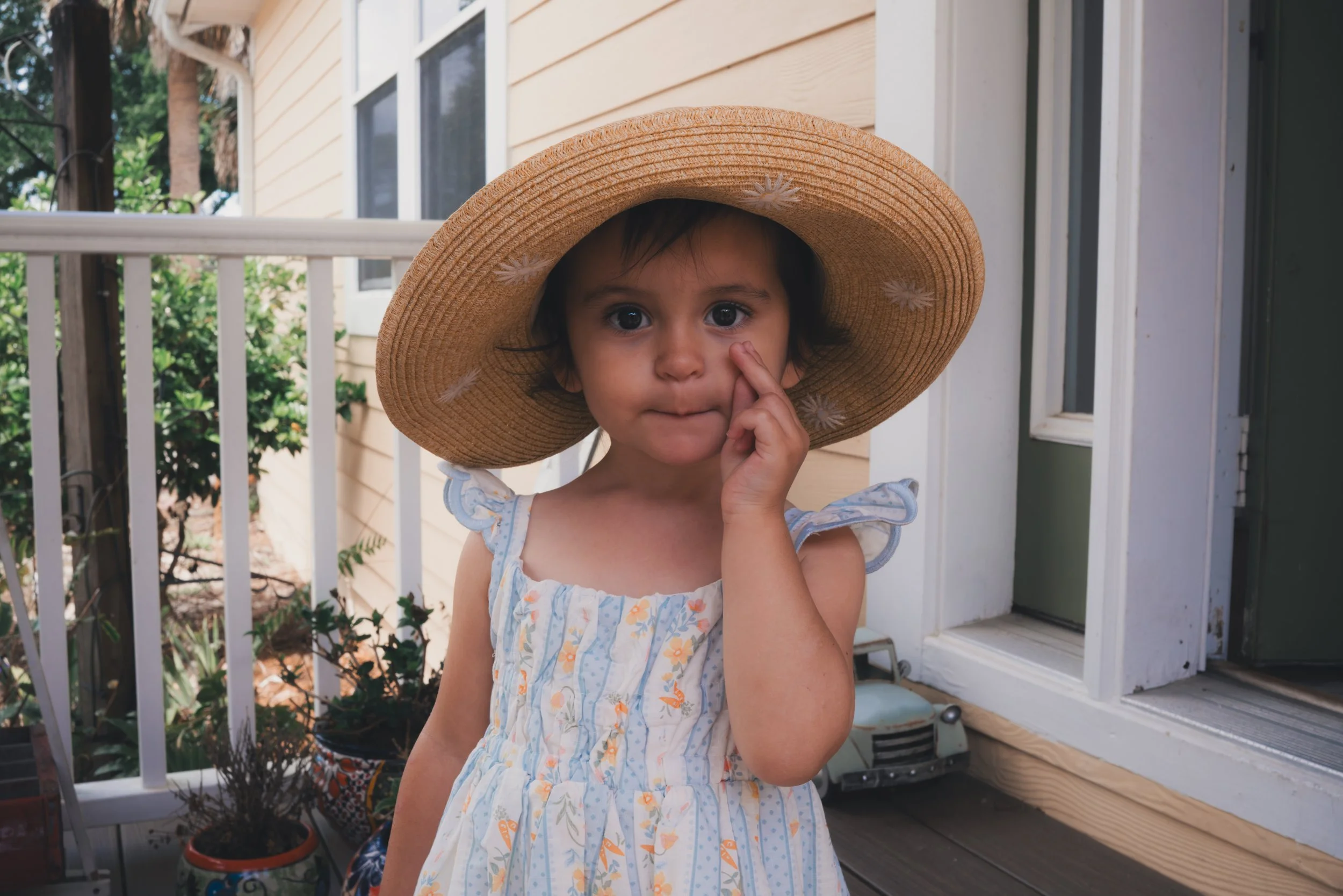 Young girl with big eyes wearing a large straw hat and a sleeveless dress with a floral pattern, standing in front of a house with a porch, plants, and a white railing.