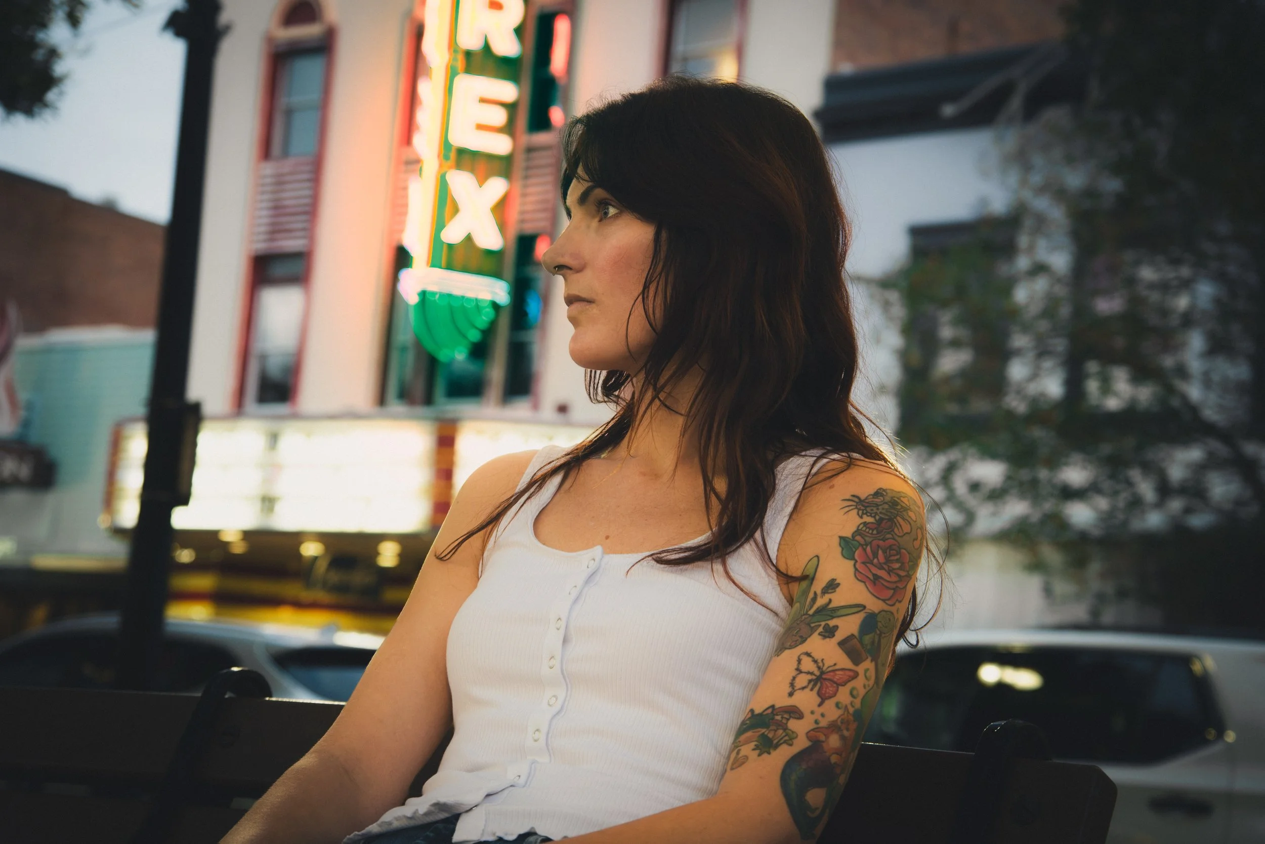 A woman with dark hair and colorful tattoos on her arm sitting in a car at dusk, with a neon sign reading 'FIRE EX' in the background.