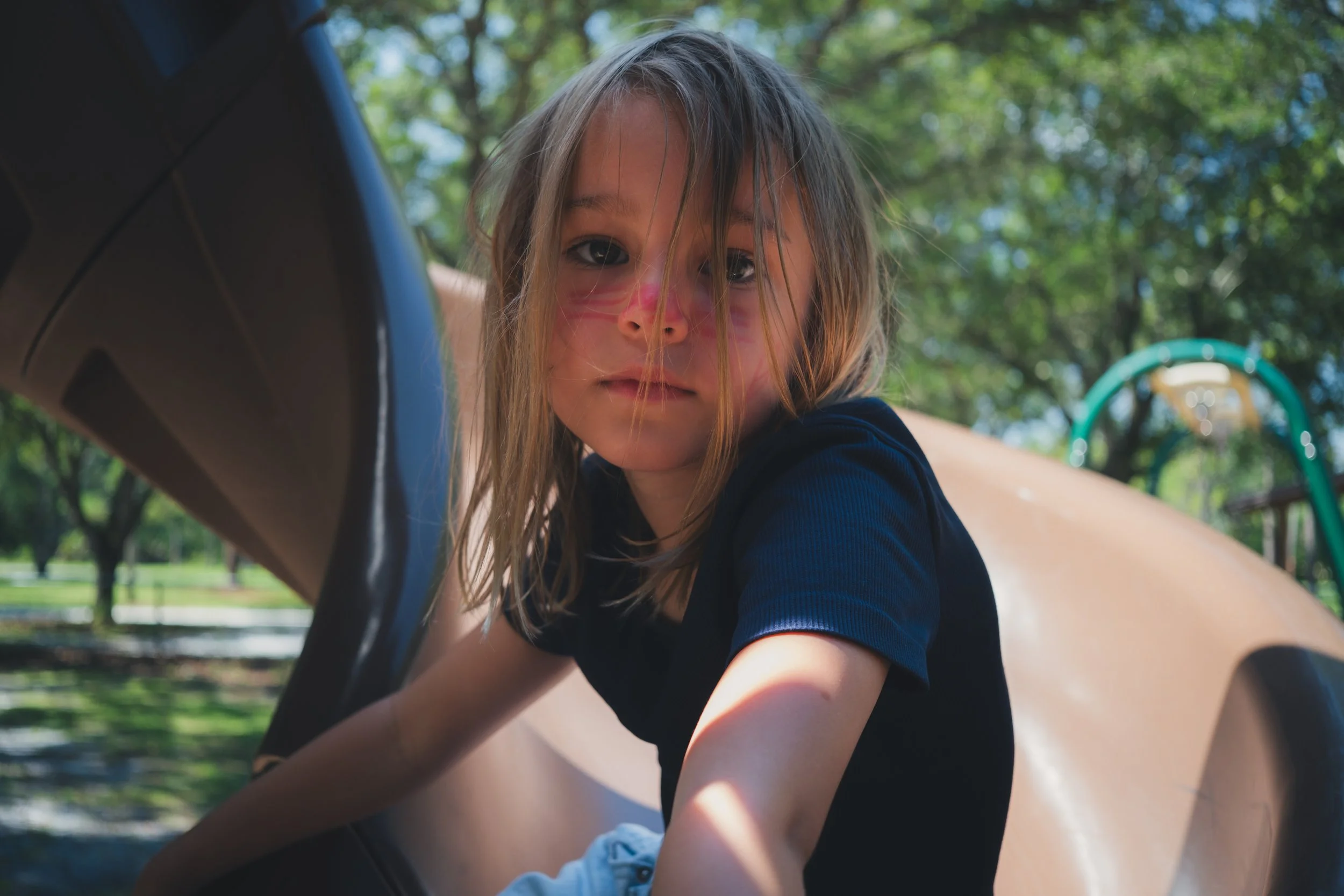 A young girl with long hair and face paint, lying on a playground slide in a park with green trees in the background.