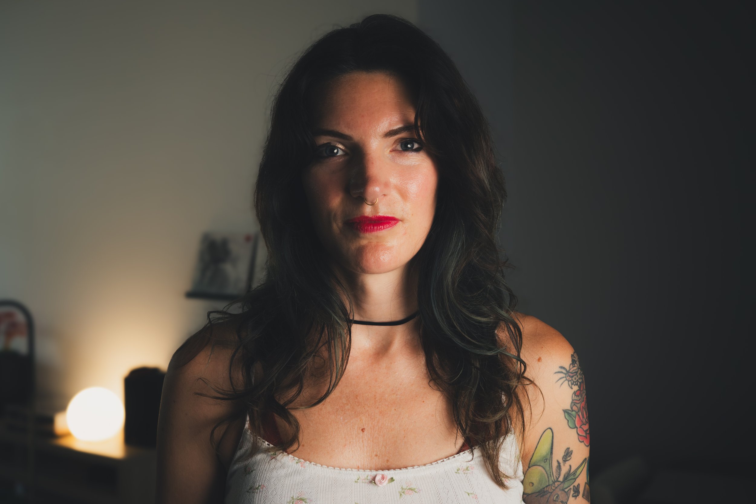 Portrait of a woman with dark wavy hair, tattoos on her right arm, wearing a choker necklace and a floral white tank top, with a softly lit background including a lamp and blurred decor.