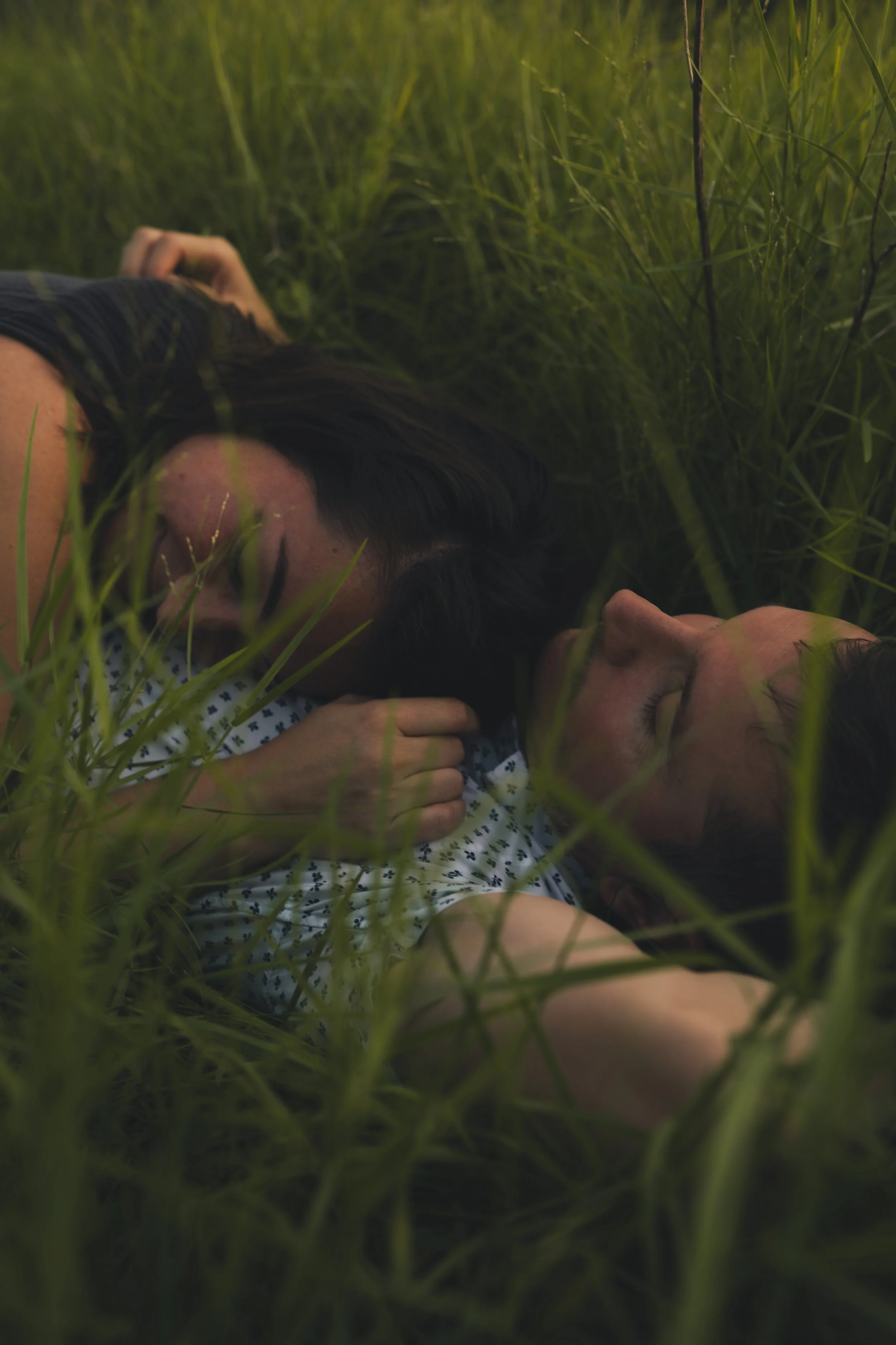 Two people lying down in tall green grass with their eyes closed, appearing peaceful and relaxed.