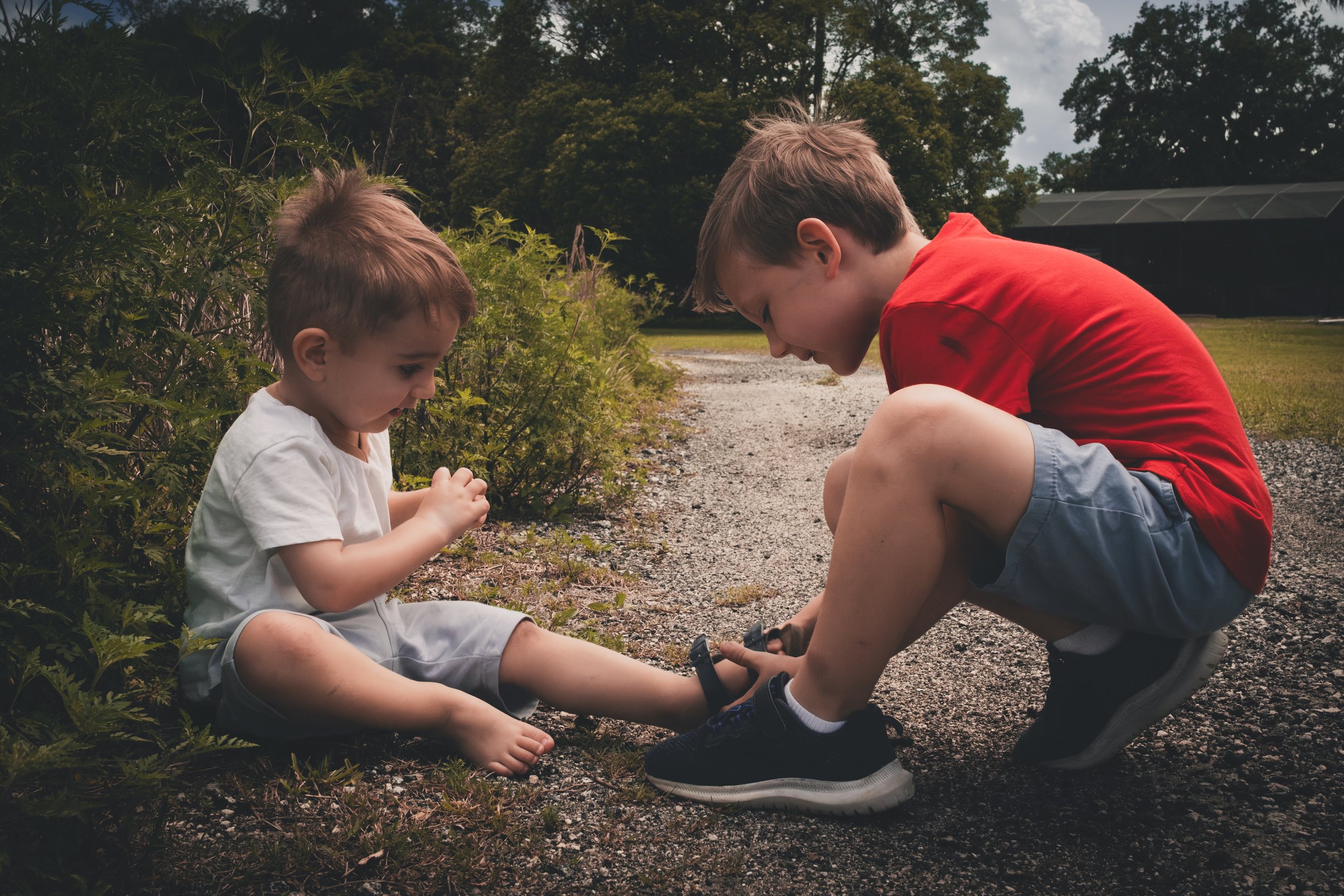 Two young boys sitting on a gravel path outdoors, one is helping the other lace up his shoes, with green bushes and trees in the background under a cloudy sky.
