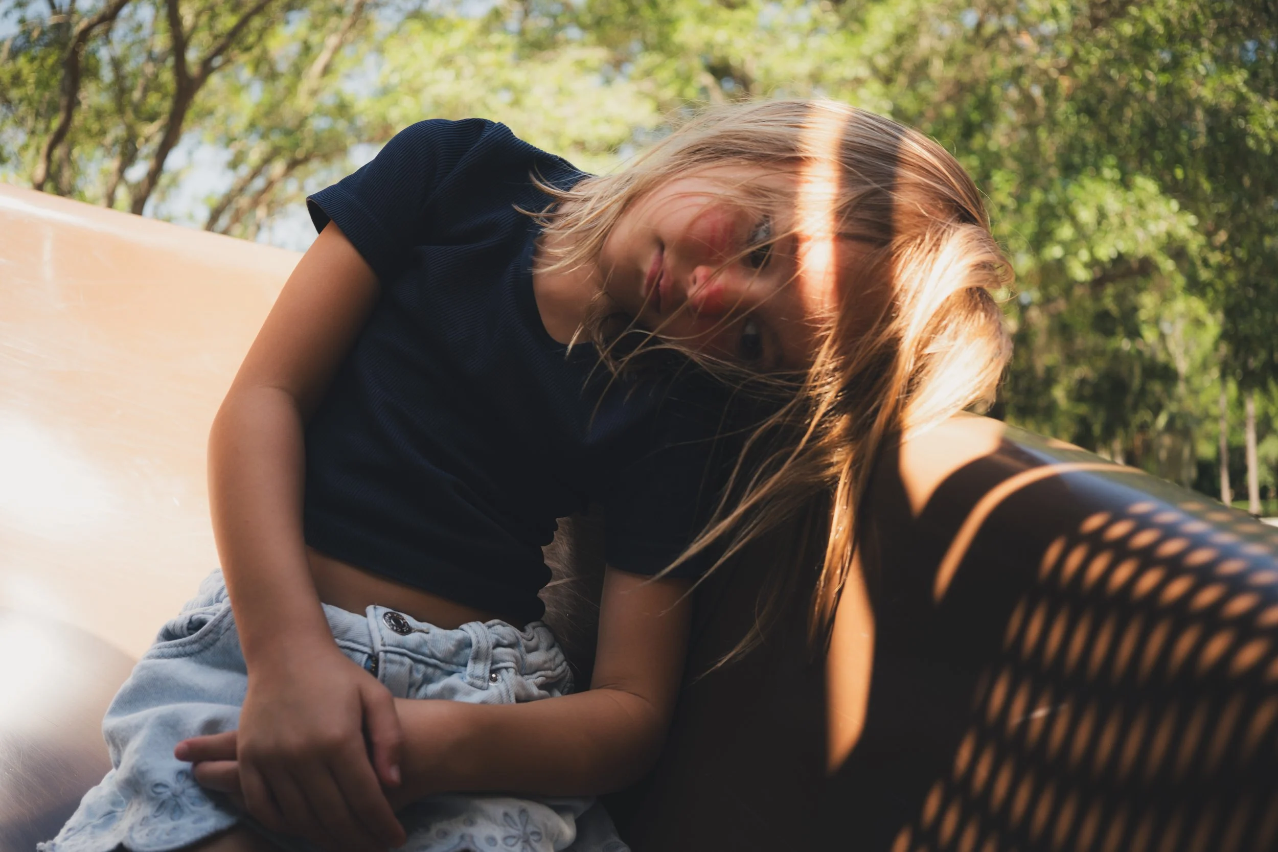 A young girl with long blonde hair, wearing a black shirt and light blue denim shorts, leans against a skateboard in an outdoor park with trees and greenery in the background, sunlight filtering through the leaves.