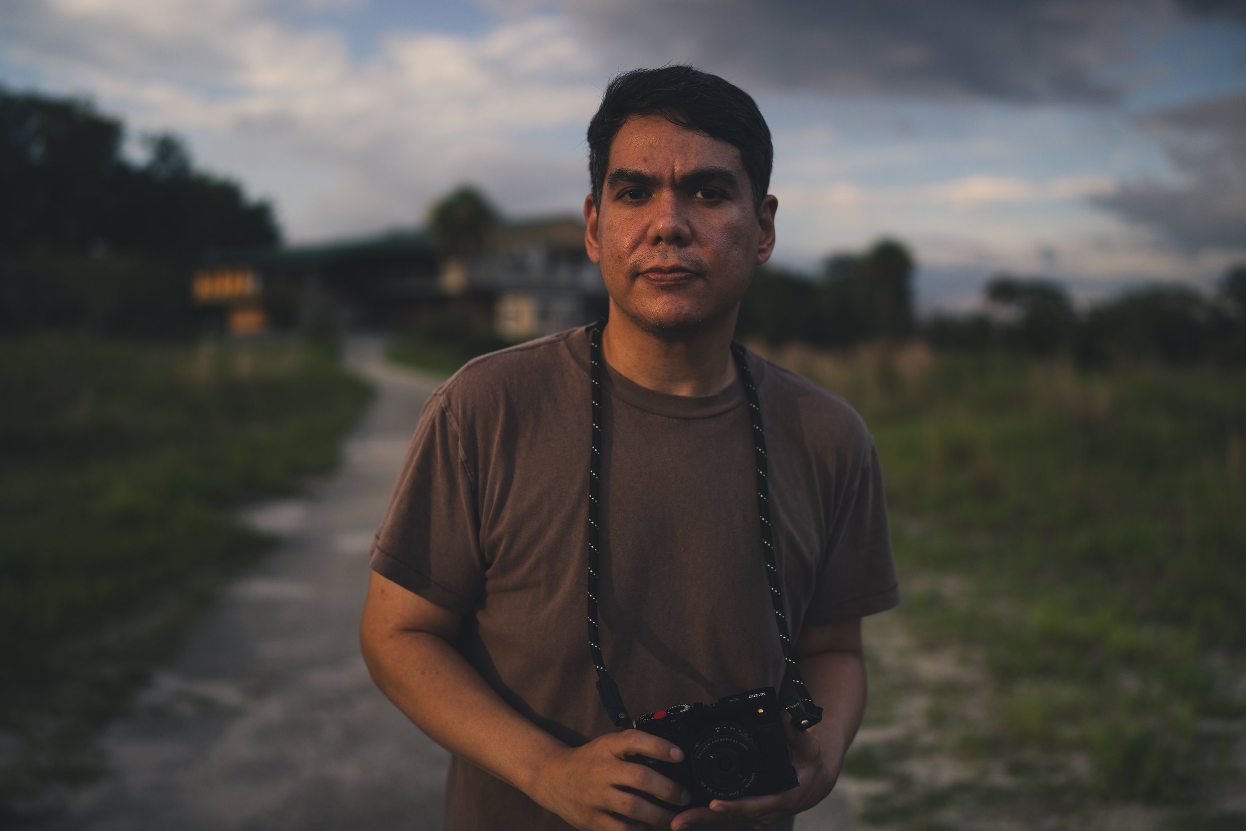 A man with short dark hair and a camera hanging around his neck stands outdoors on a trail with a cloudy sky in the background.