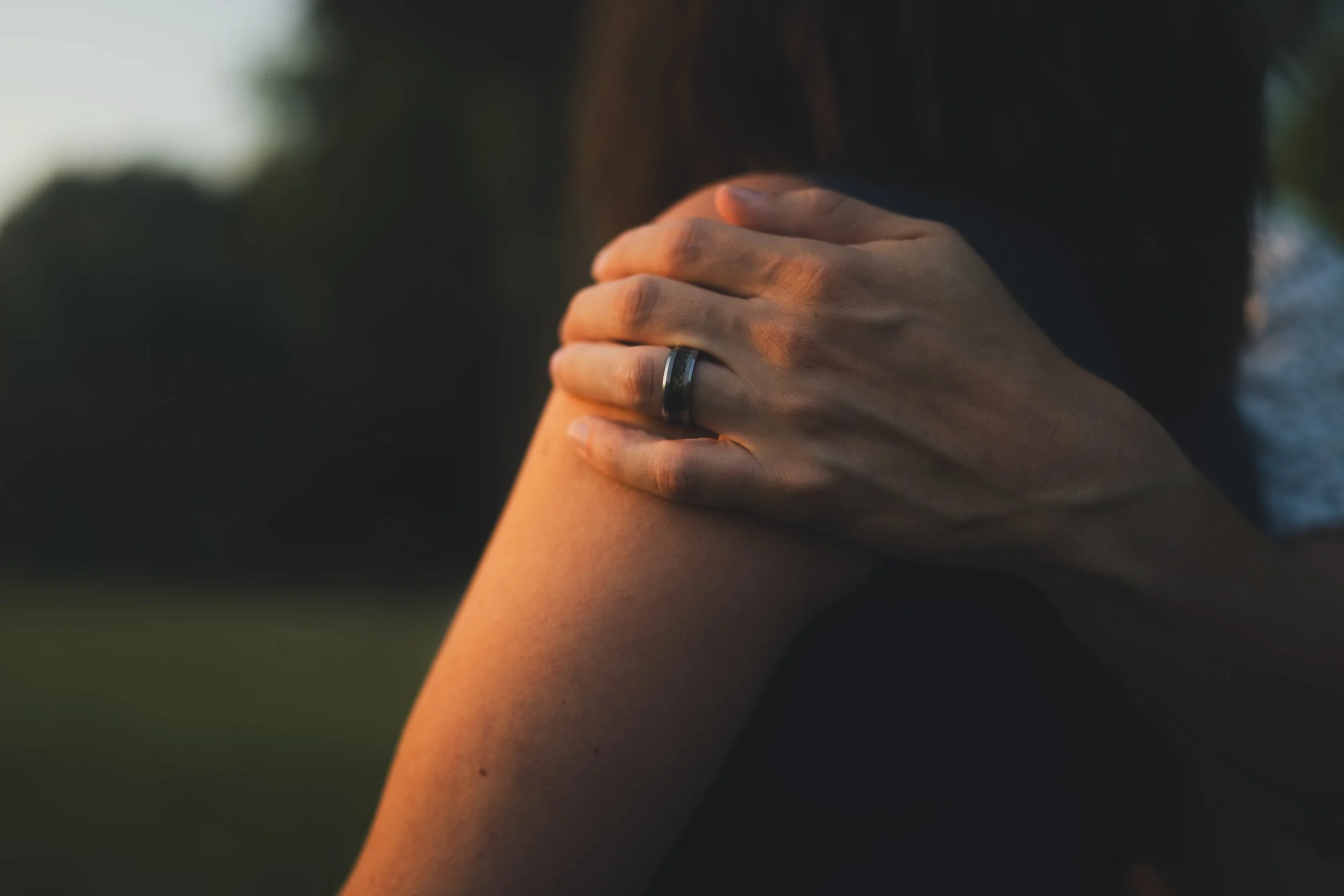 A person holding their hand over their face with fingers touching their forehead, wearing a black wedding ring, with a dark background and sunset lighting.