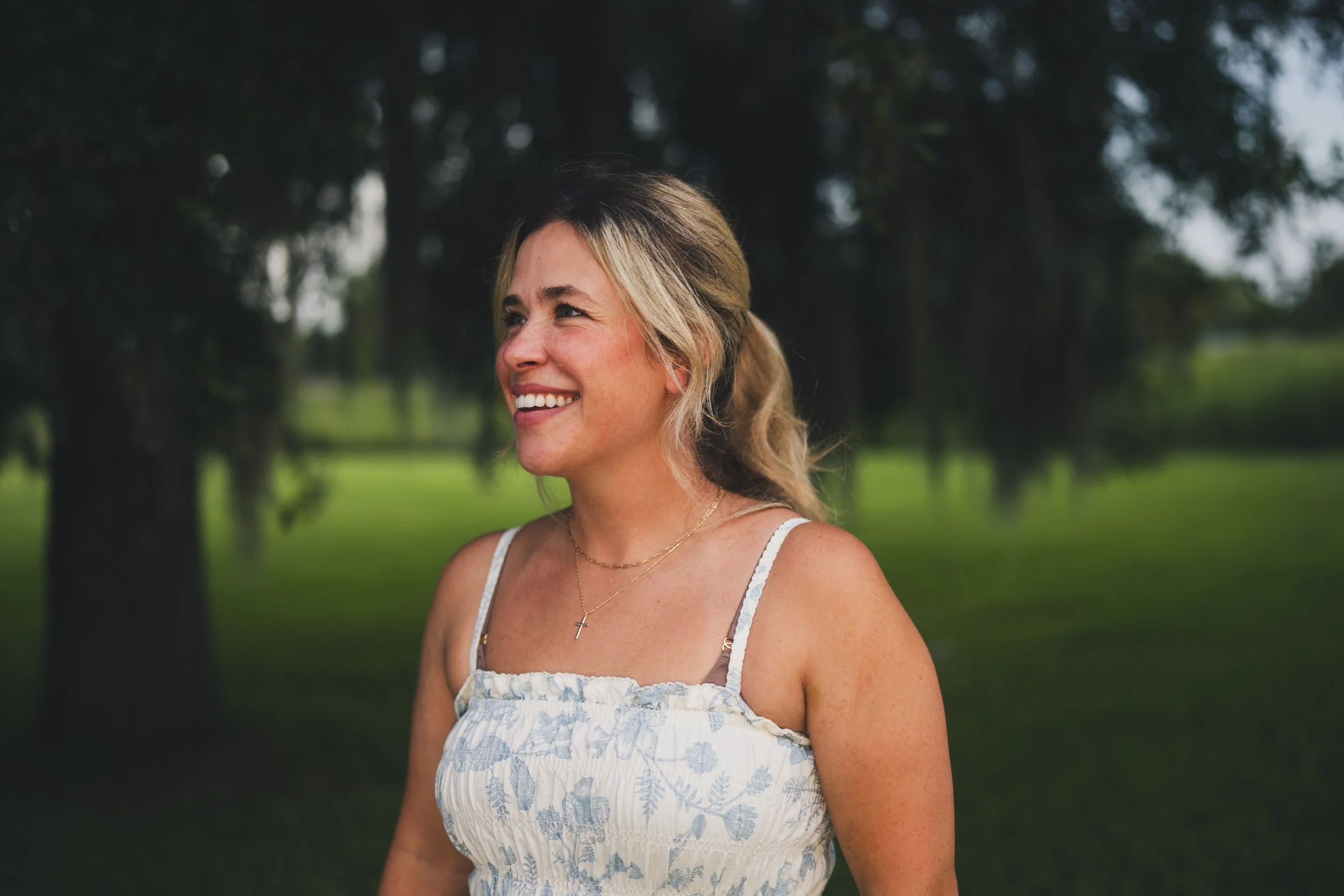 A woman with blonde hair smiling outdoors in front of trees, wearing a white and blue patterned sundress and a gold cross necklace.