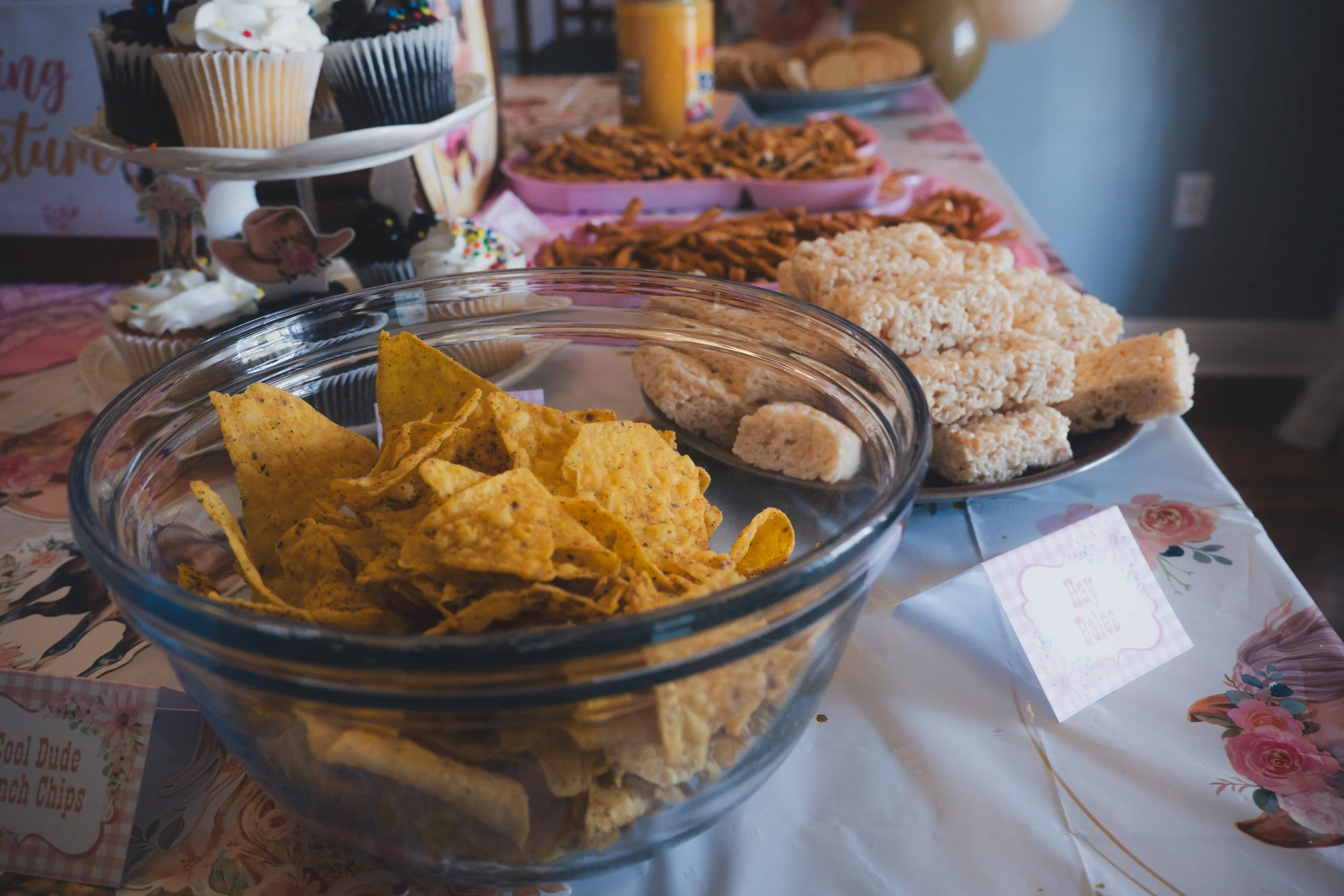 A glass bowl of seasoned tortilla chips in the foreground, with an assortment of other snacks and baked goods on a table, including rice Krispies treats, cookies, and cupcakes with white frosting and colorful sprinkles.