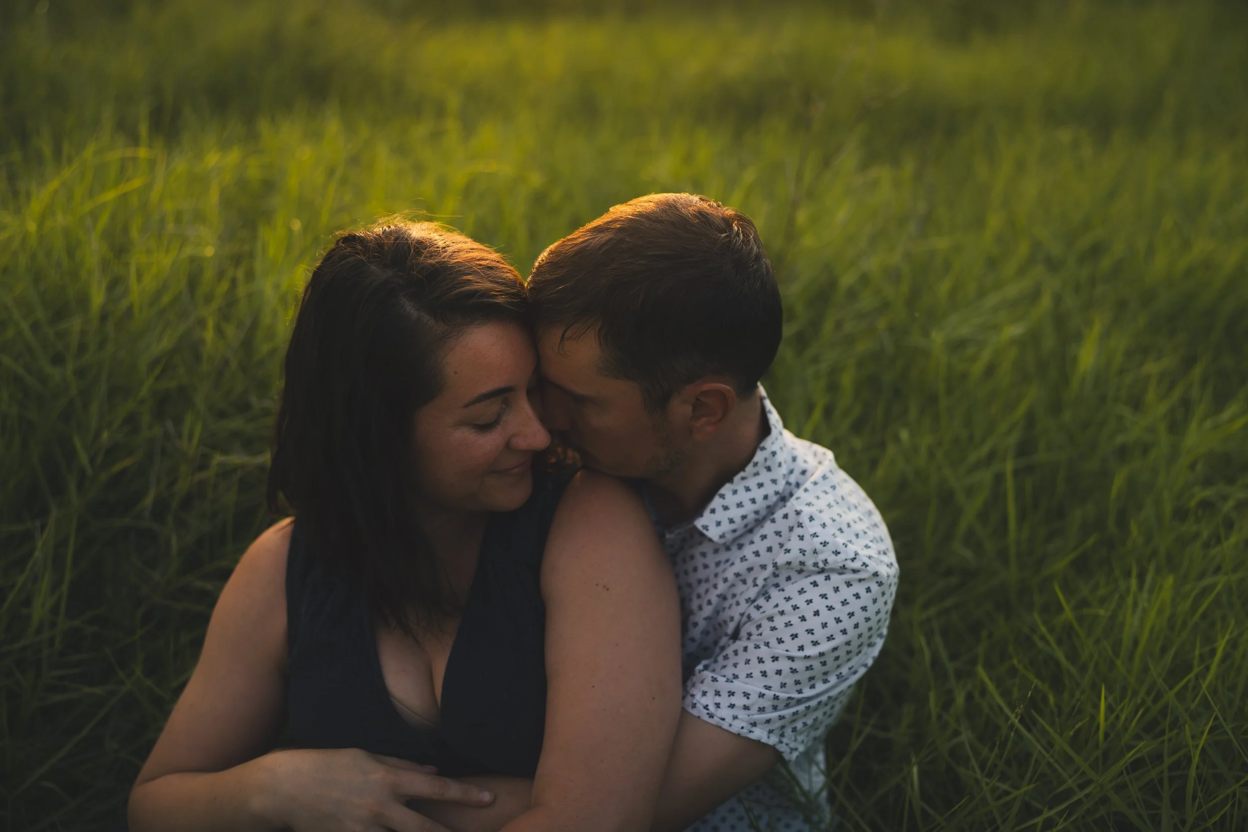 A couple embracing each other in a grassy field during sunset, with their foreheads touching and eyes closed.