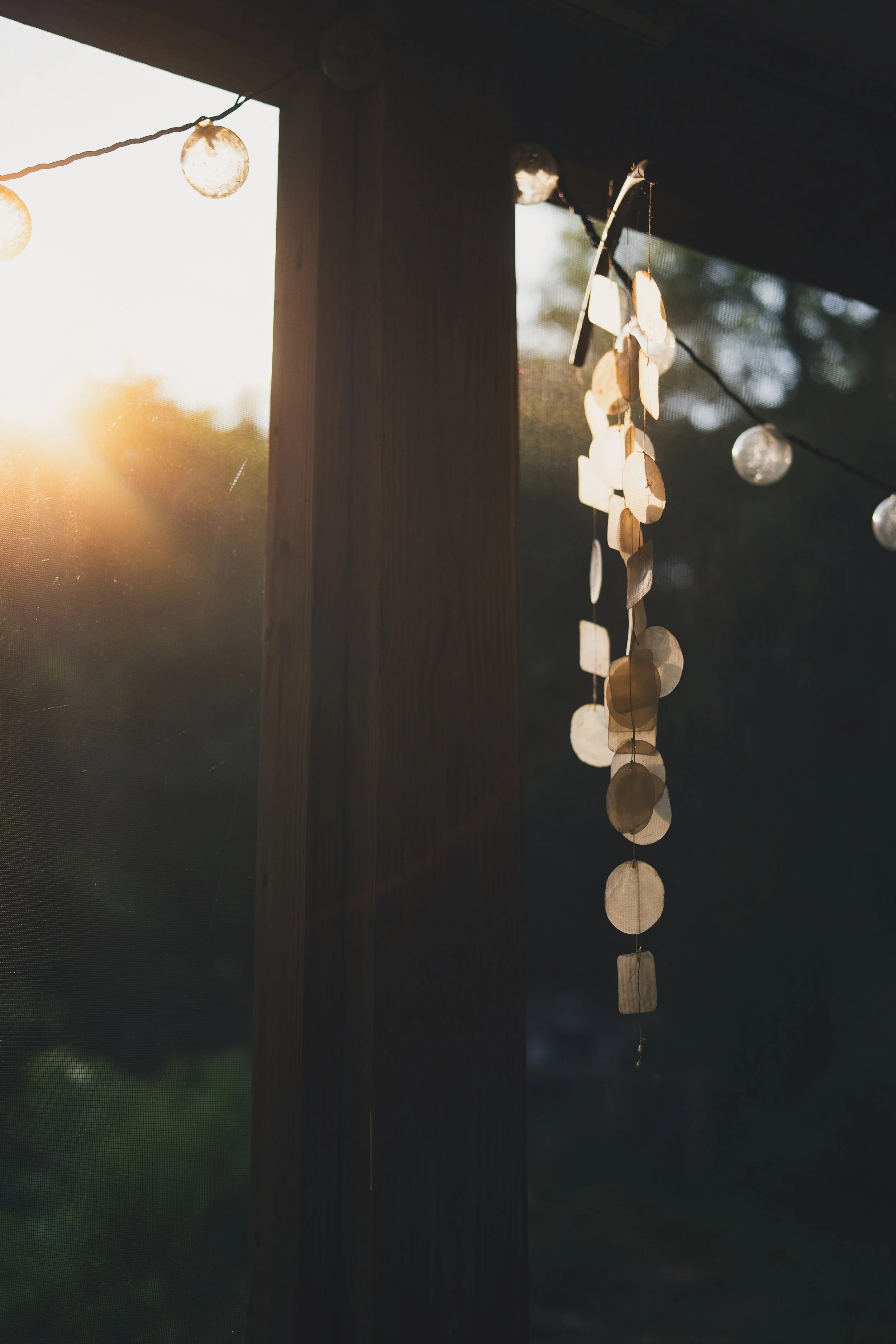 Close-up of decorative hanging wind chimes made of circular and rectangular paper or wood pieces, hanging from a string on a house's porch, with sunlight filtering through and a blurred outdoor background.