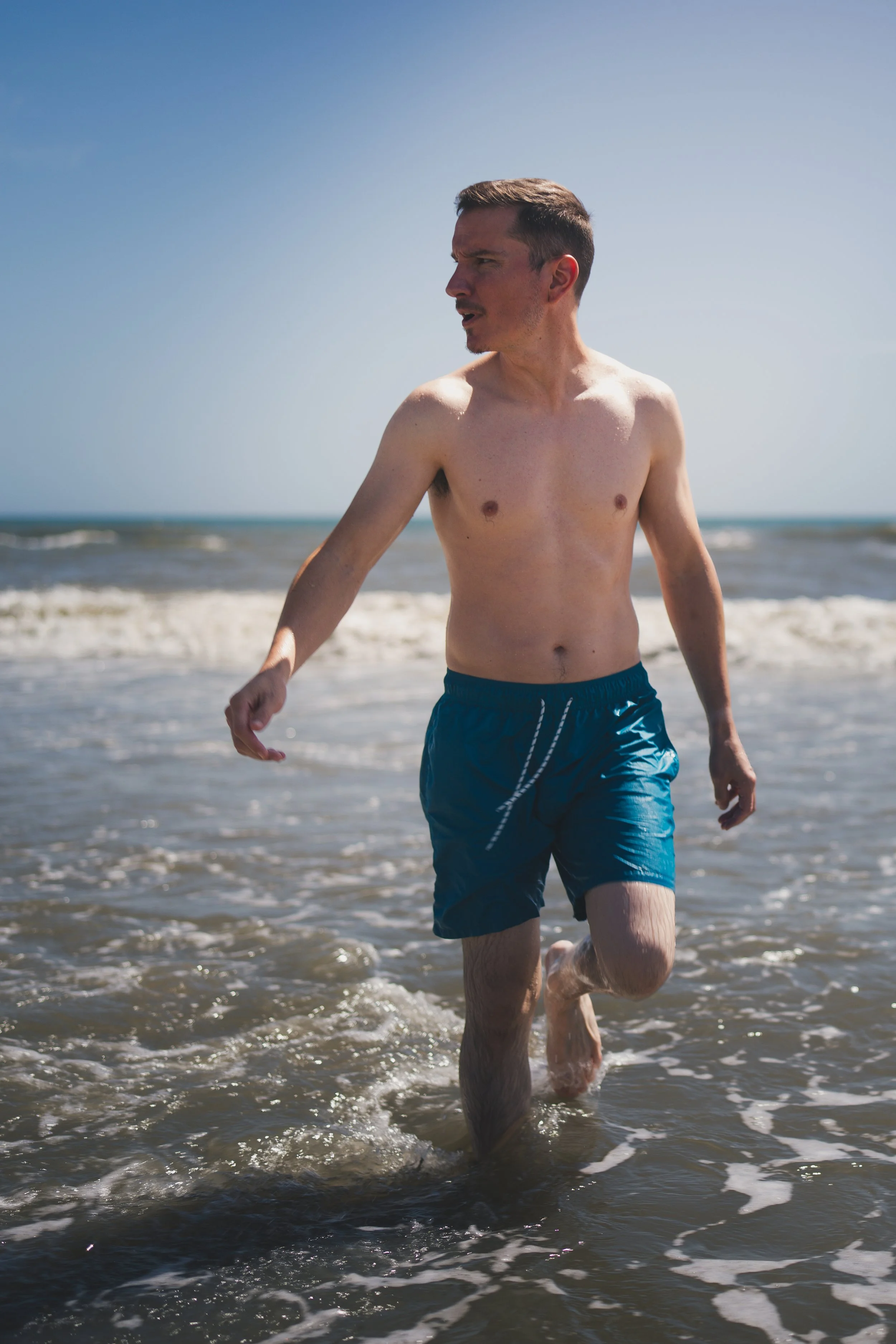A young man in blue swim trunks running into the ocean, with waves splashing around his legs and a clear blue sky in the background.