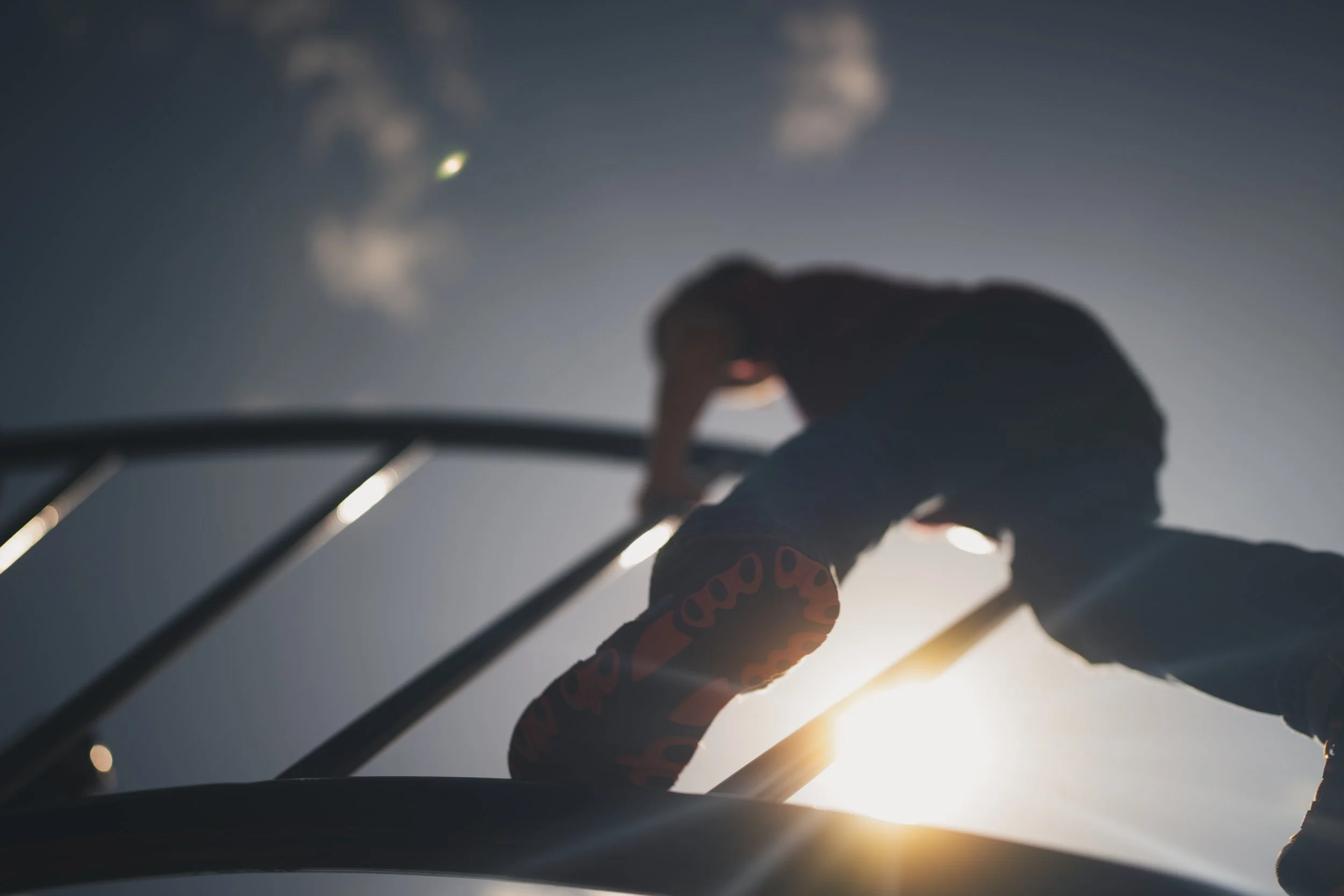 A person reaching upward with a hand, with the sun low in the sky creating a bright light and soft focus effect.