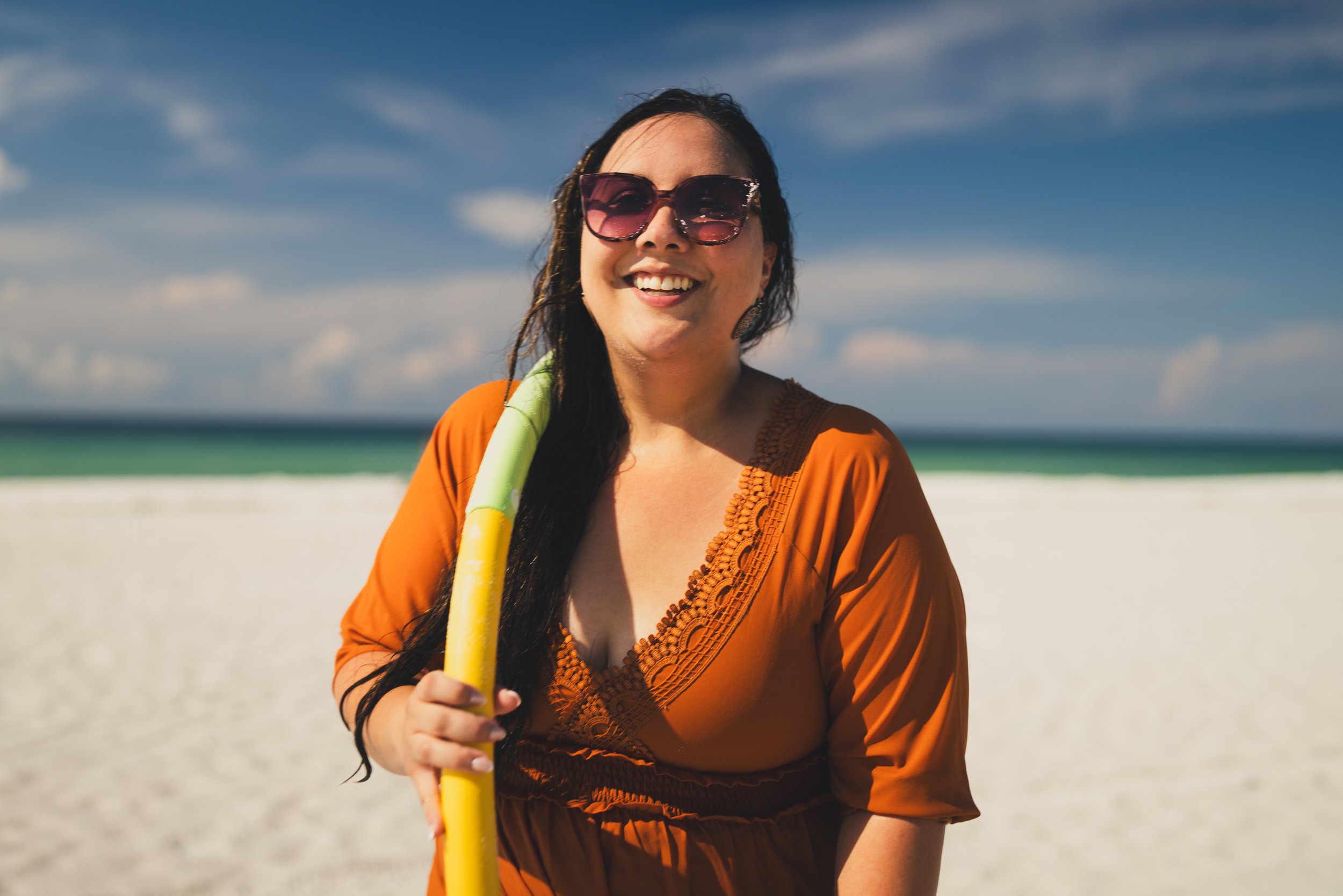 A woman with long dark hair, wearing sunglasses and an orange dress, smiling while holding a yellow and green snorkel on a beach with blue sky and ocean in the background.
