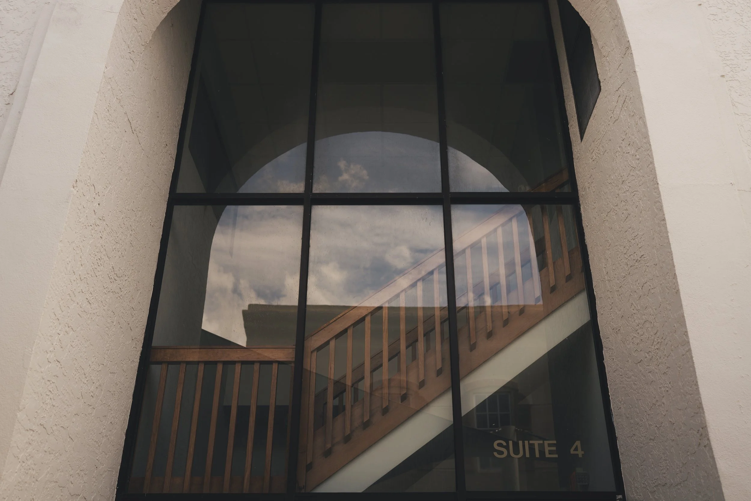 Building with a large glass window reflecting the sky and clouds, beige textured exterior wall, and a sign indicating 'Suite 4'.