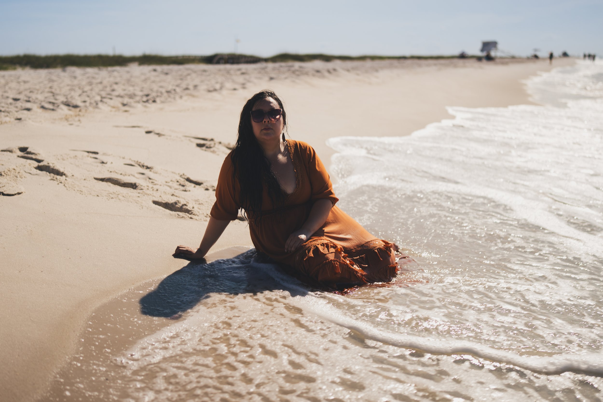 A woman in sunglasses and an orange dress sitting on the sandy beach near the water's edge, with a few people and structures in the background.