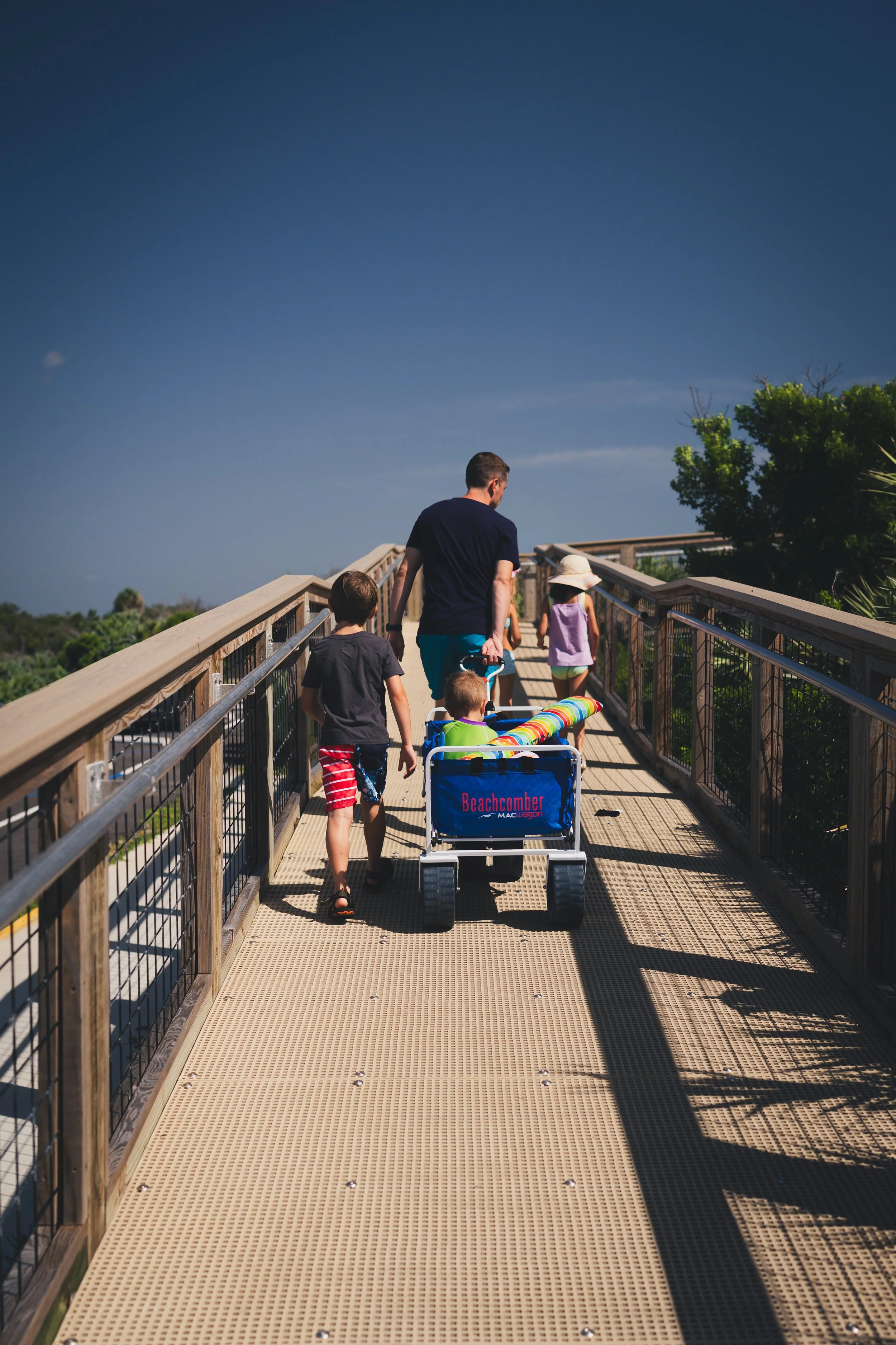 A man and three children walk along a raised wooden and metal walkway outdoors on a sunny day. The man pulls a beach cart labeled 'Beachcomber' with a colorful striped umbrella inside. The children wear casual summer clothes, and one girl has a wide-