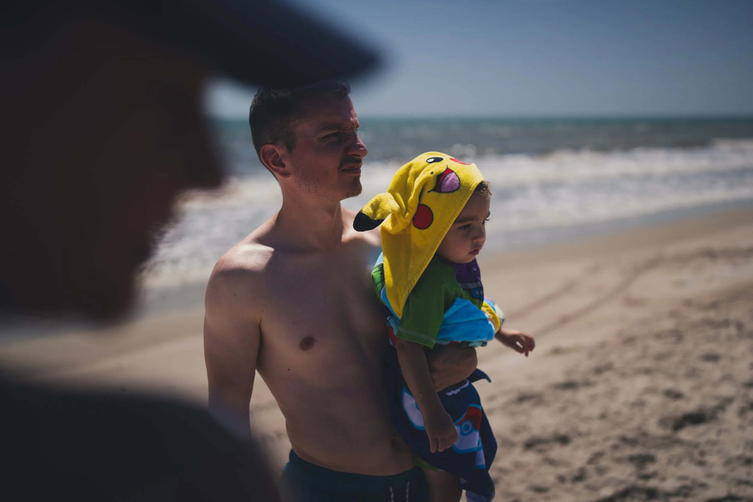 A man holding a young boy at the beach, with the man shirtless and the boy wearing a Pikachu towel hood.