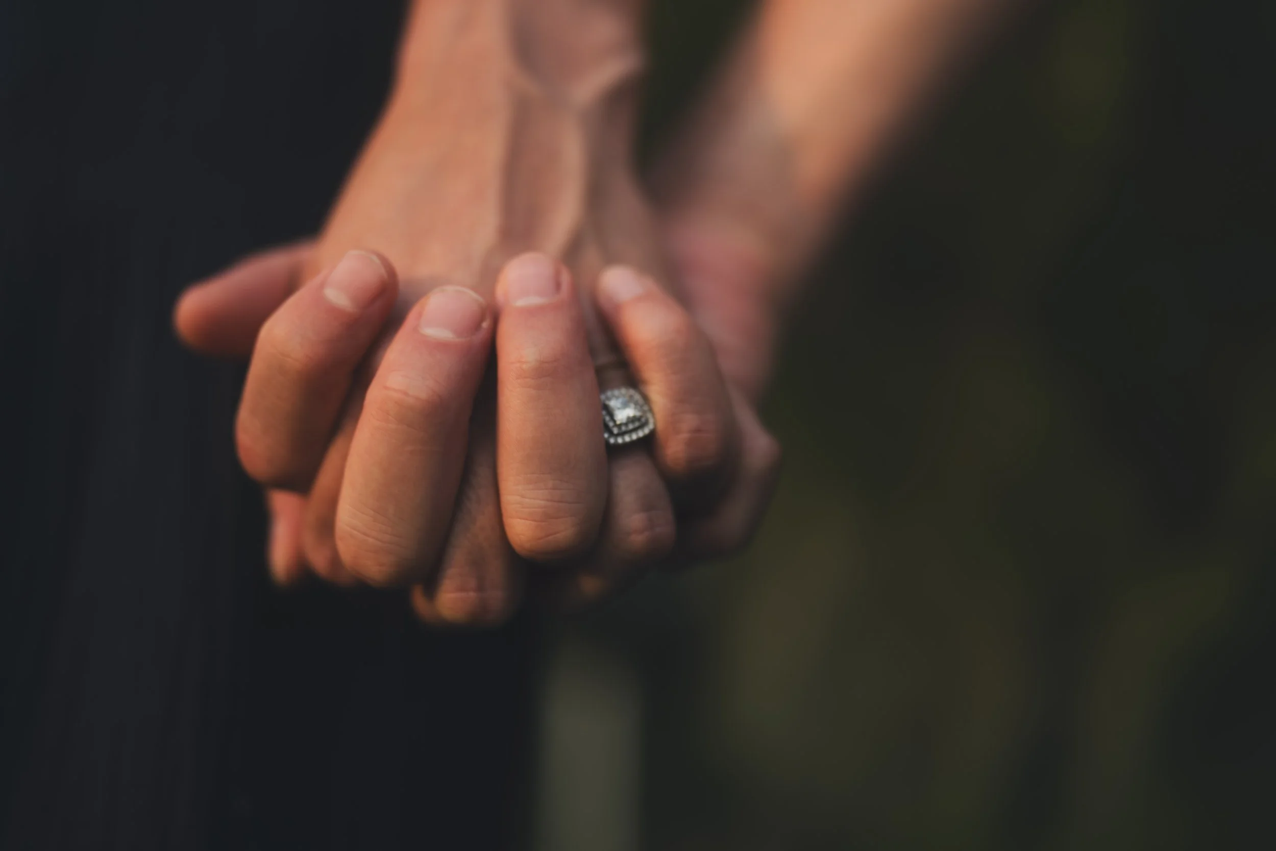 Close-up of a couple holding hands, with the woman's hand showing an engagement ring, outdoors at night.