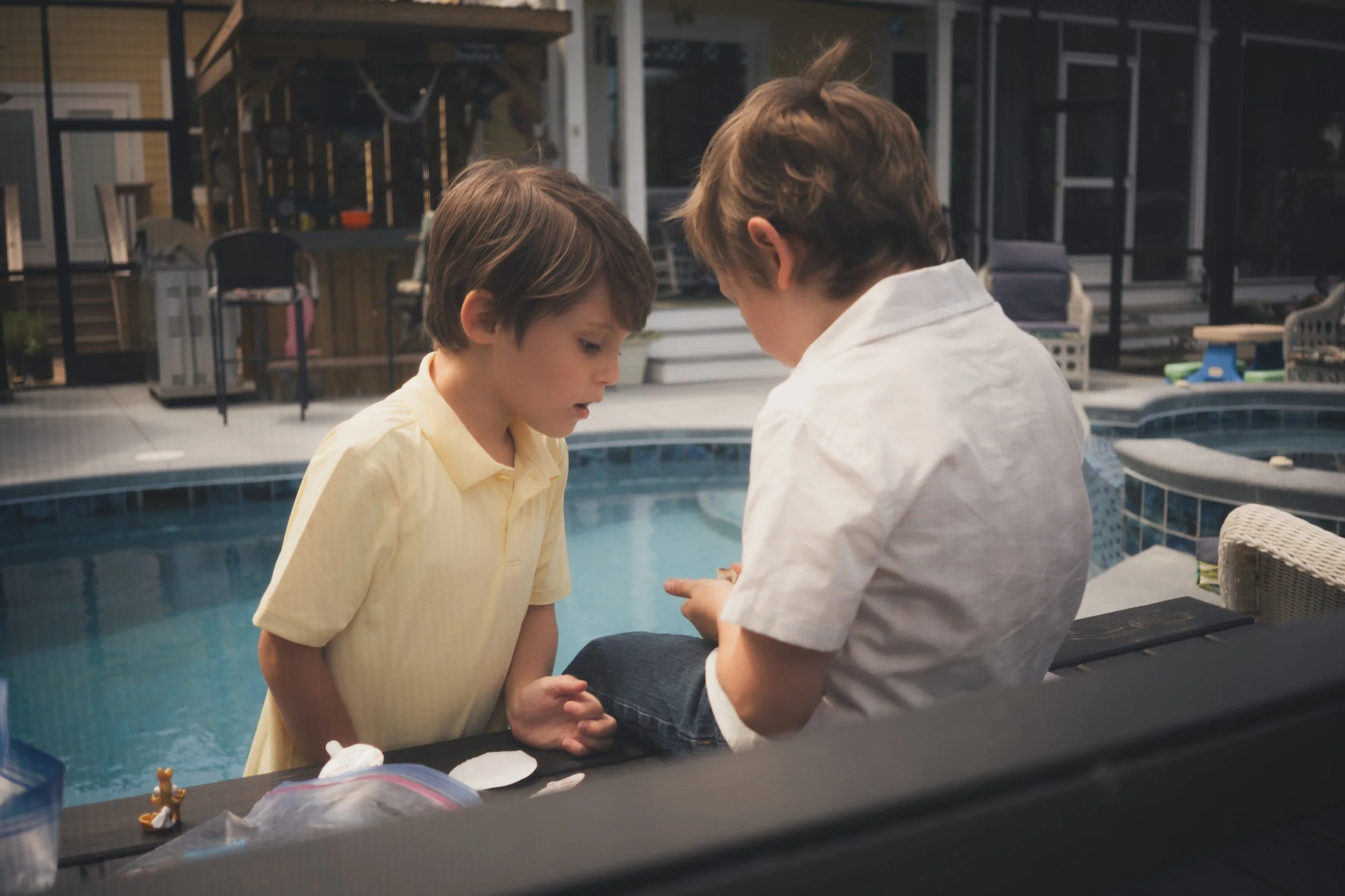 Two young boys are arm wrestling by a swimming pool, with one boy wearing a yellow shirt and the other wearing a white shirt, in a backyard setting.