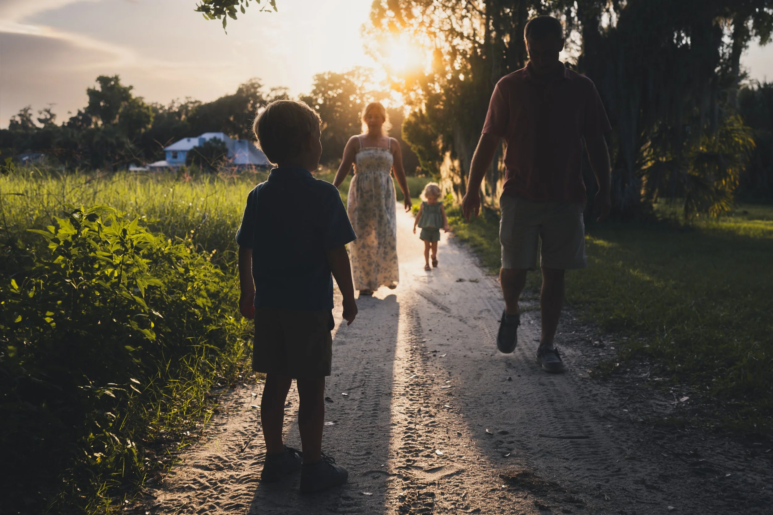 A group of five people, including children and adults, walking on a dirt path during sunset with trees and houses in the background.