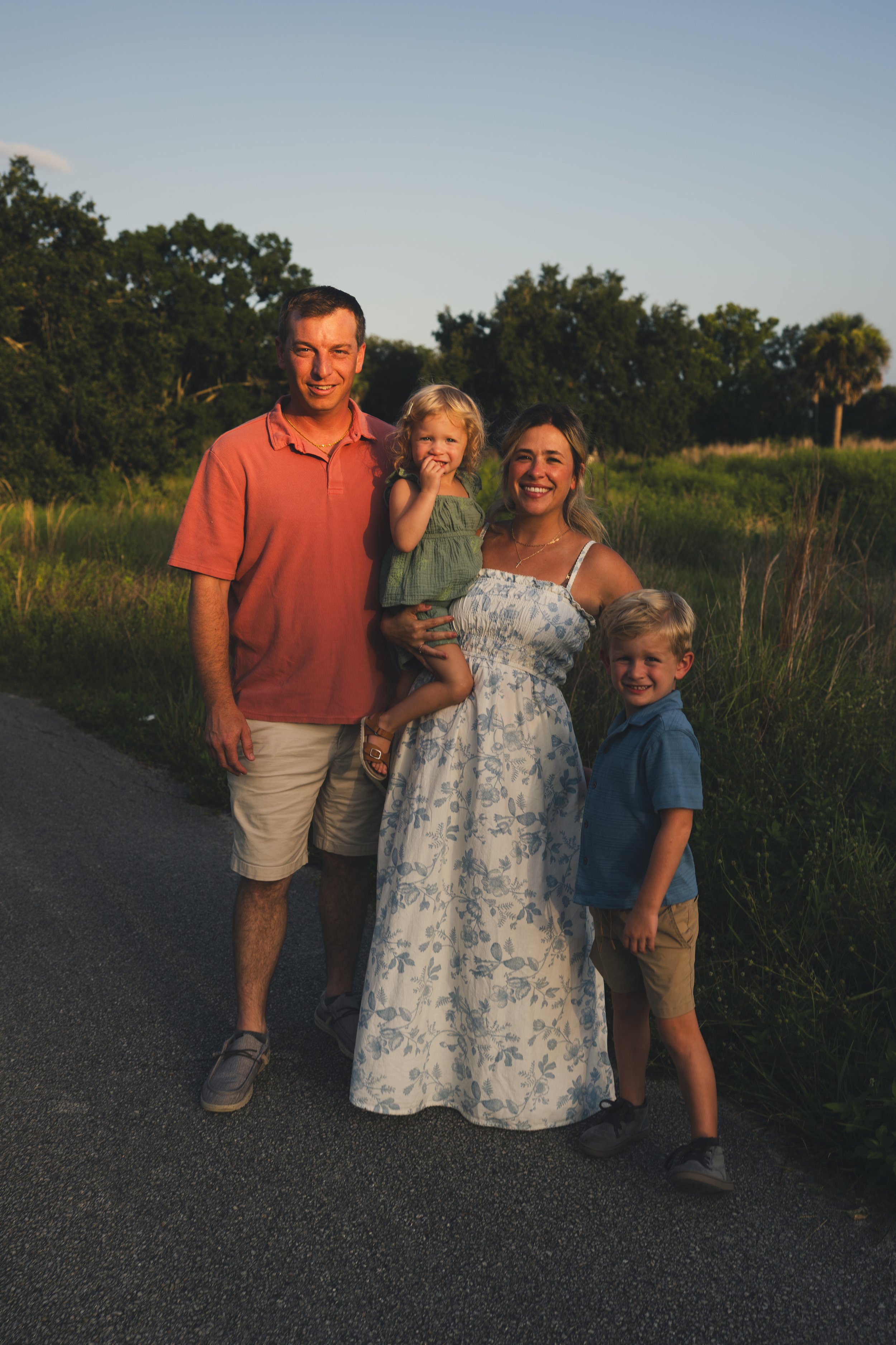A family of four outdoors during sunset, smiling, with a man, a woman holding a young girl, and a boy standing beside her, in a grassy area with trees in the background.