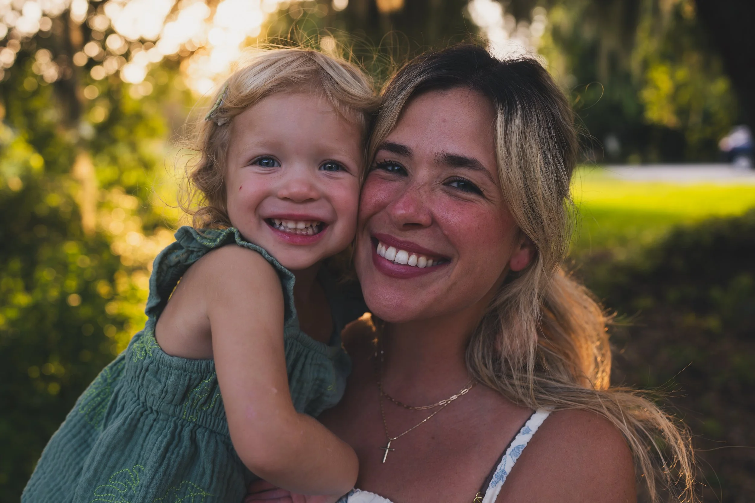 A woman and a young girl smiling and hugging each other outdoors with blurred trees and sunlight in the background.