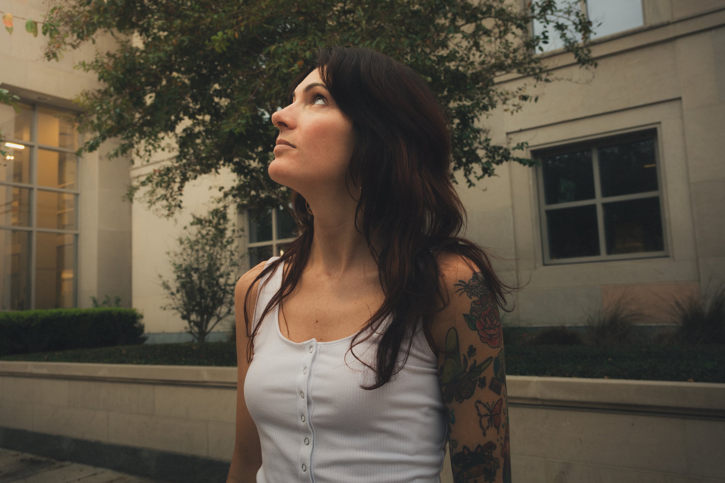 A woman with dark hair and tattoos on her arm wearing a white tank top, looking up outdoors near buildings and trees.