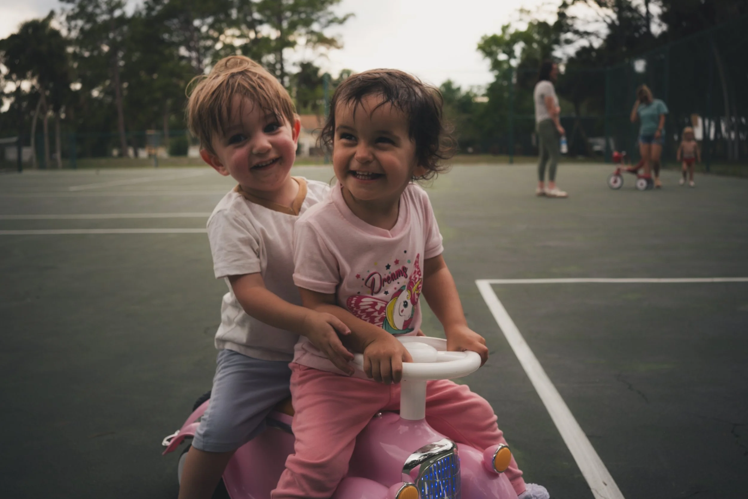 Two young children, a boy and a girl, sitting on a pink ride-on toy motorcycle on a tennis court, smiling and enjoying their time. In the background, there are adults and another child, with trees surrounding the court, indicating a park or recreatio