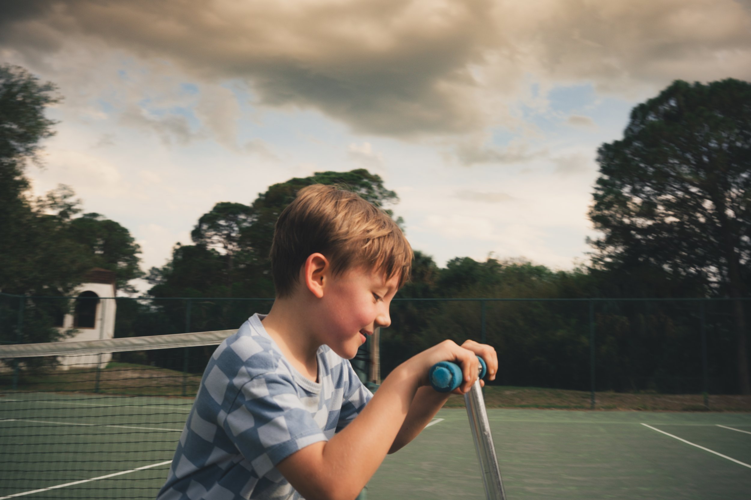 A young boy with light brown hair and wearing a blue and white striped shirt riding a scooter on a tennis court, with a cloudy sky and trees in the background.