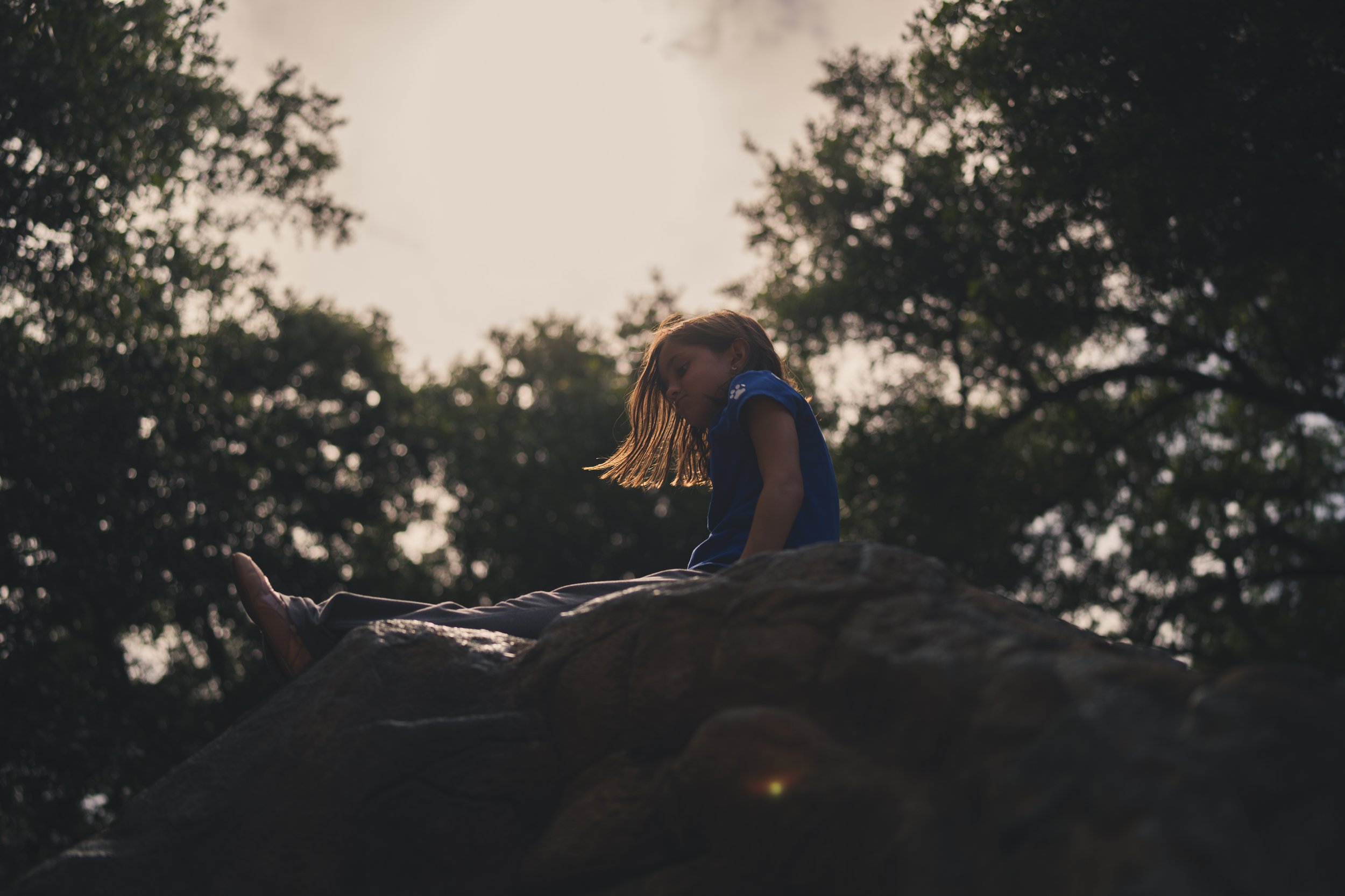 A young girl with long hair sitting on a large rock outdoors, with trees and a cloudy sky in the background.