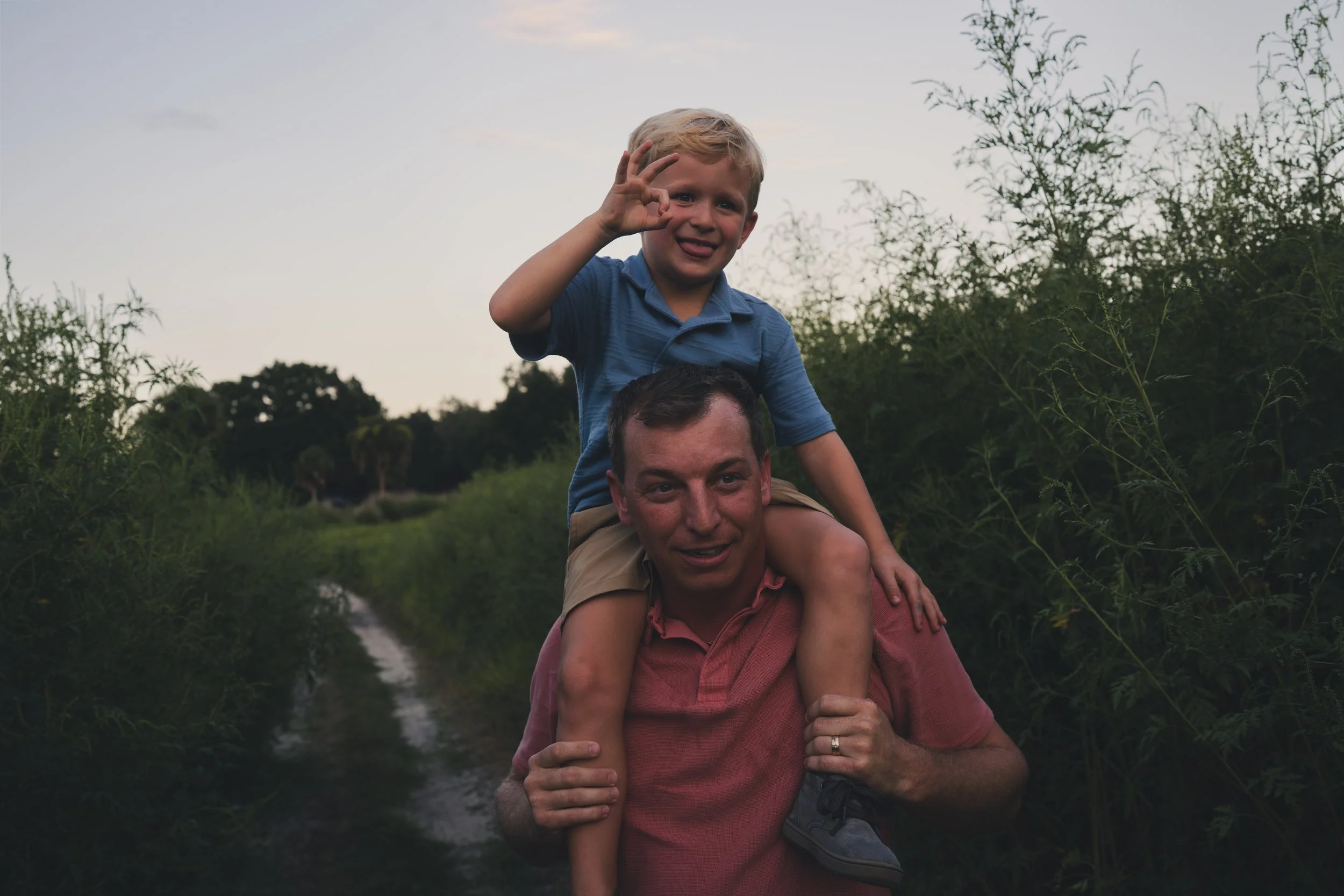 A man carrying a young boy on his shoulders through a lush outdoor setting, both smiling and enjoying the moment during sunset or dusk.