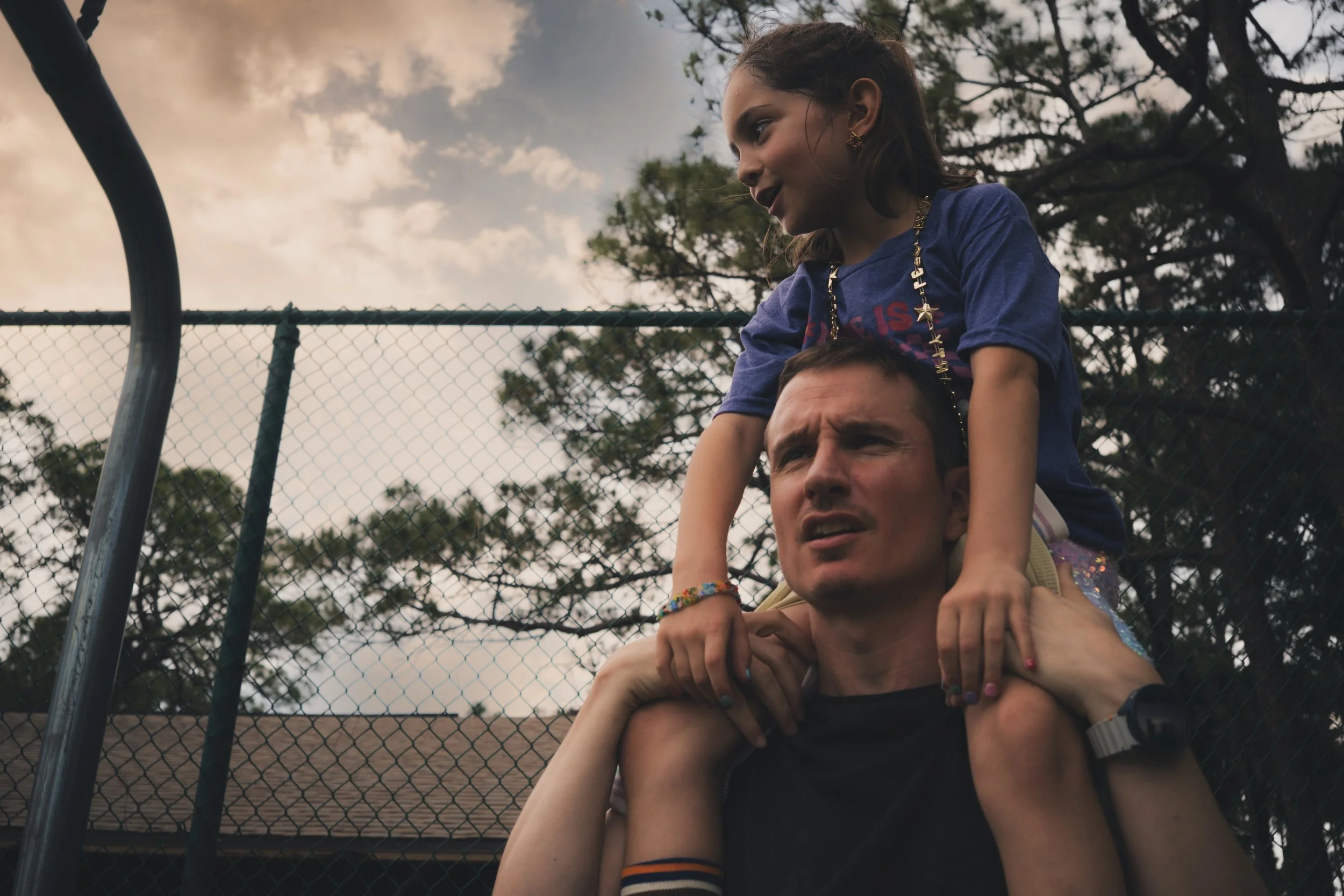A young girl sitting on a man's shoulders at a baseball field, with a chain-link fence and trees in the background, during cloudy weather.