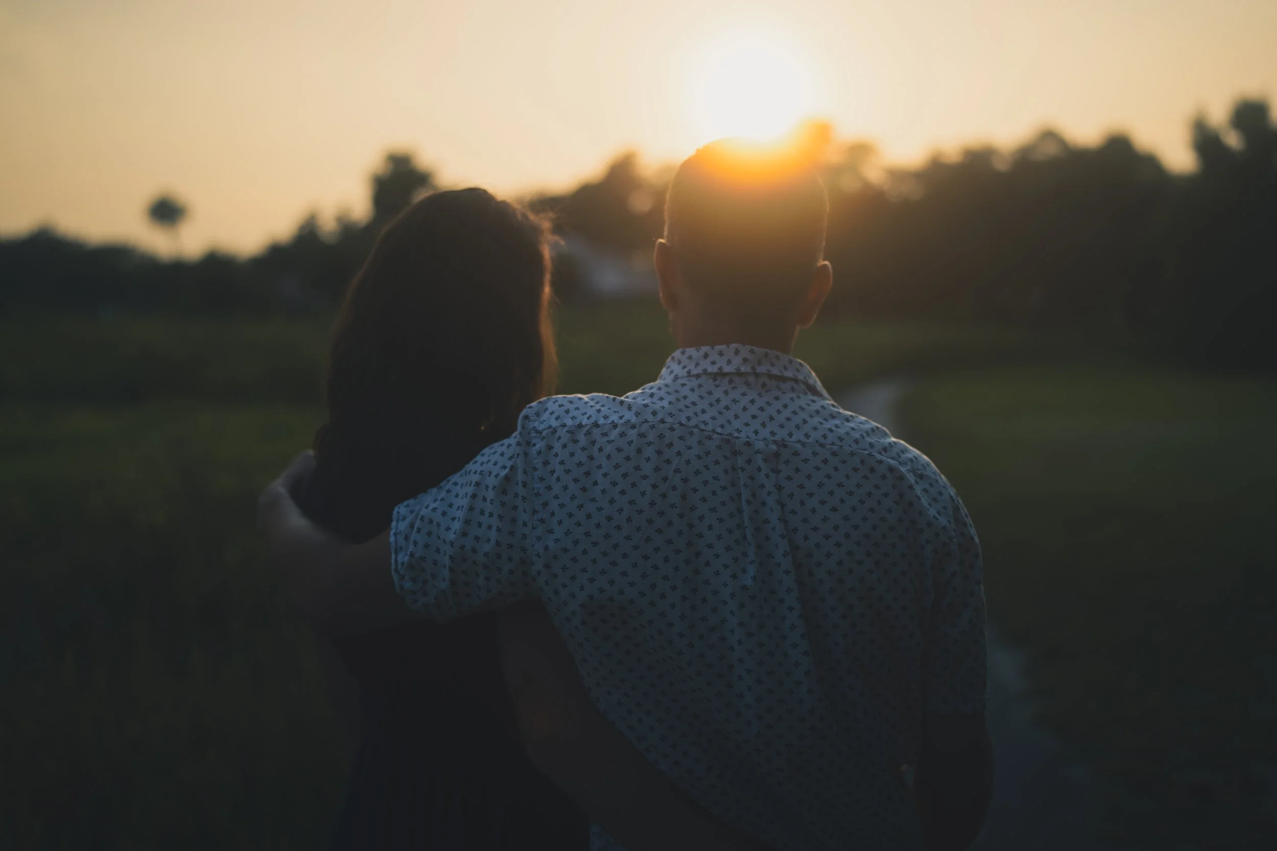 Two people are sitting together outdoors at sunset, with the sun low in the sky, and trees in the background.