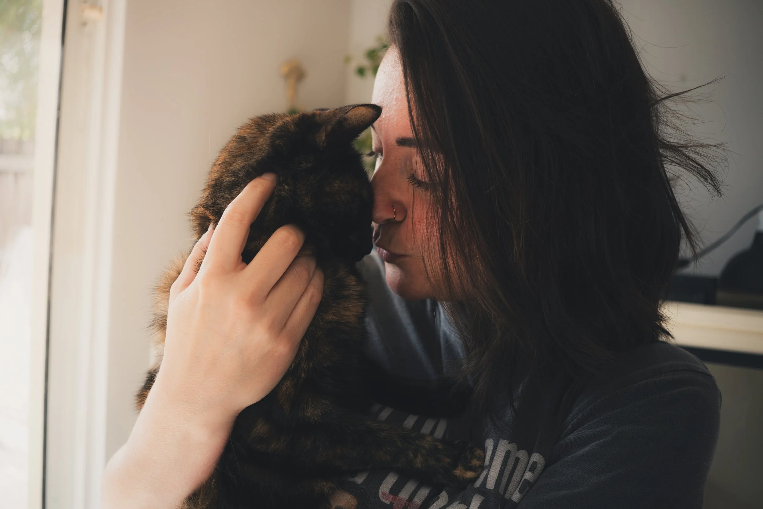 A woman with black hair and a nose piercing gently touching and leaning her forehead against a tabby cat with dark and light brown fur, both with their eyes closed, in a sunny indoor setting.