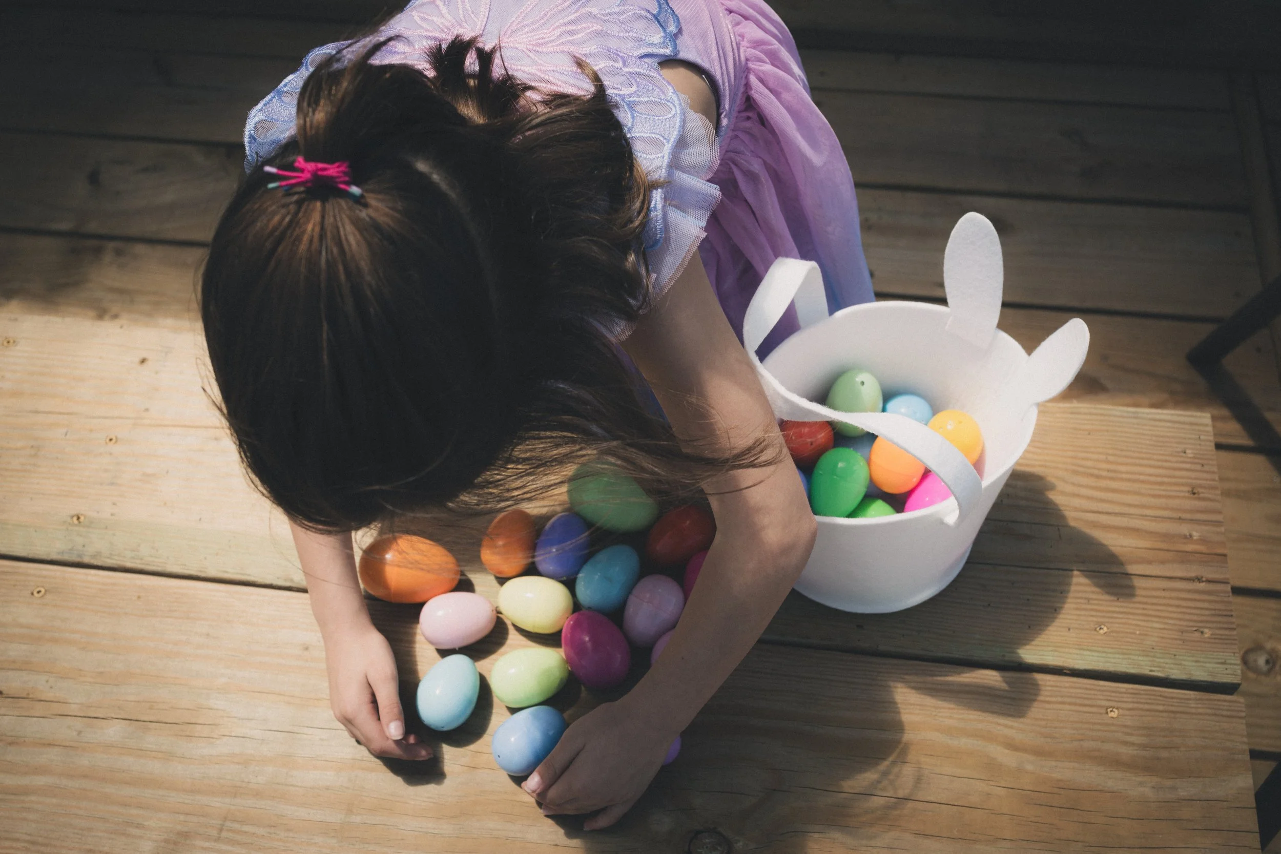 A young girl with brown hair tied with a pink hair tie, wearing a purple dress with a lace collar, is sitting on a wooden deck. She is reaching into a white container shaped like a bunny with tall ears, which is filled with colorful plastic eggs, inc