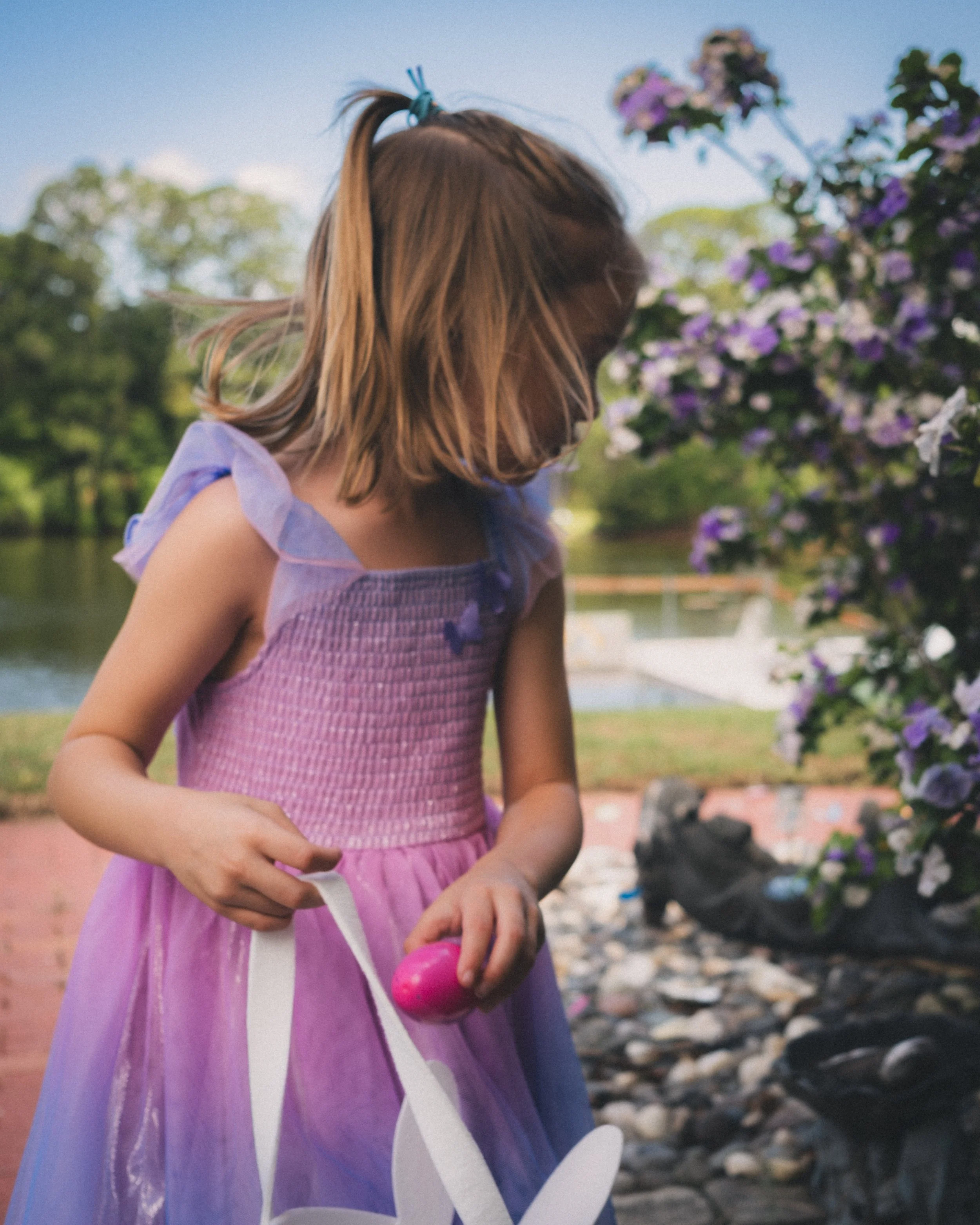 Young girl in purple sleeveless dress standing outdoors, near purple flowers, holding a pink and white toy in her hand, with a pond and trees in the background.