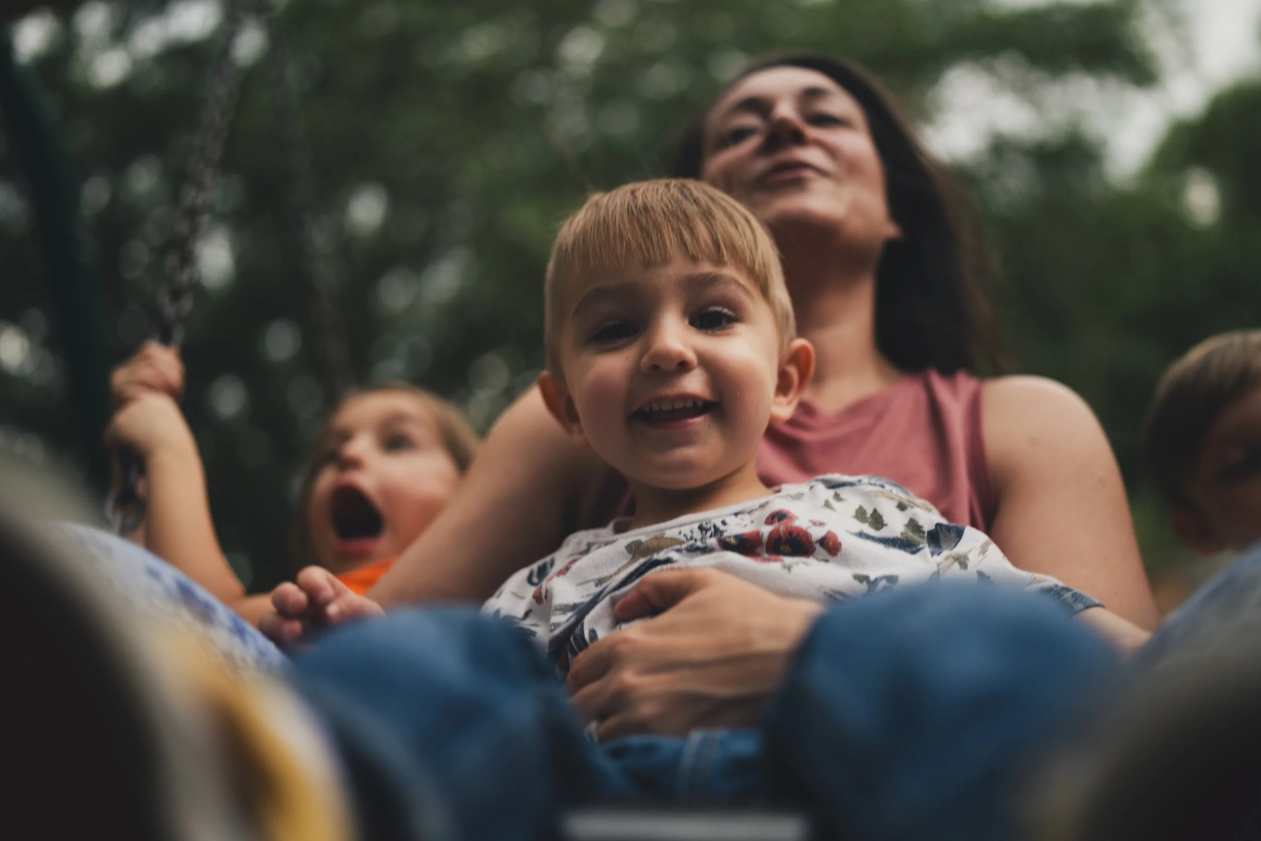 A young boy with reddish hair and a smile sitting on an adult's lap, with other children and a woman in the background, outdoors with trees and greenery.