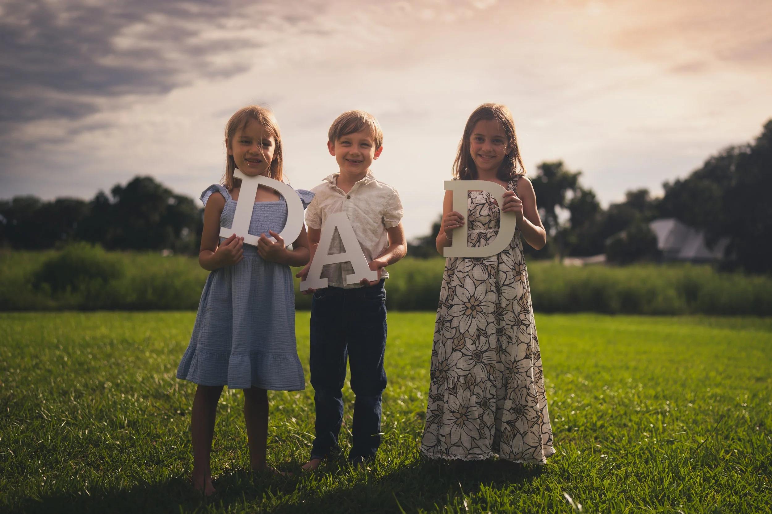 Three children, two girls and one boy, standing outdoors on grass at sunset, holding large white letters that spell 